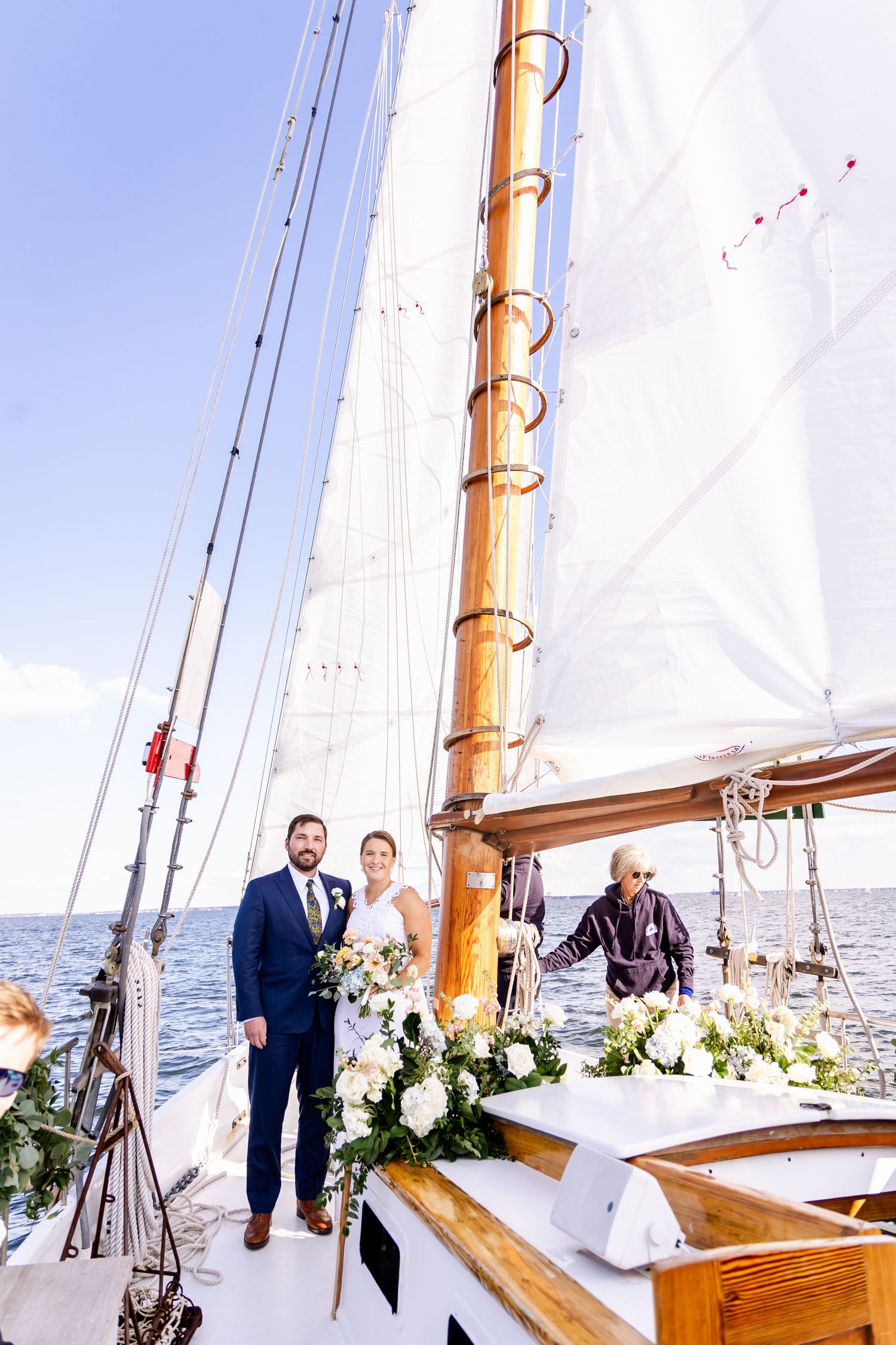 Bride and groom pose on a decorated sailboat deck with white floral arrangements and billowing sails on open water.