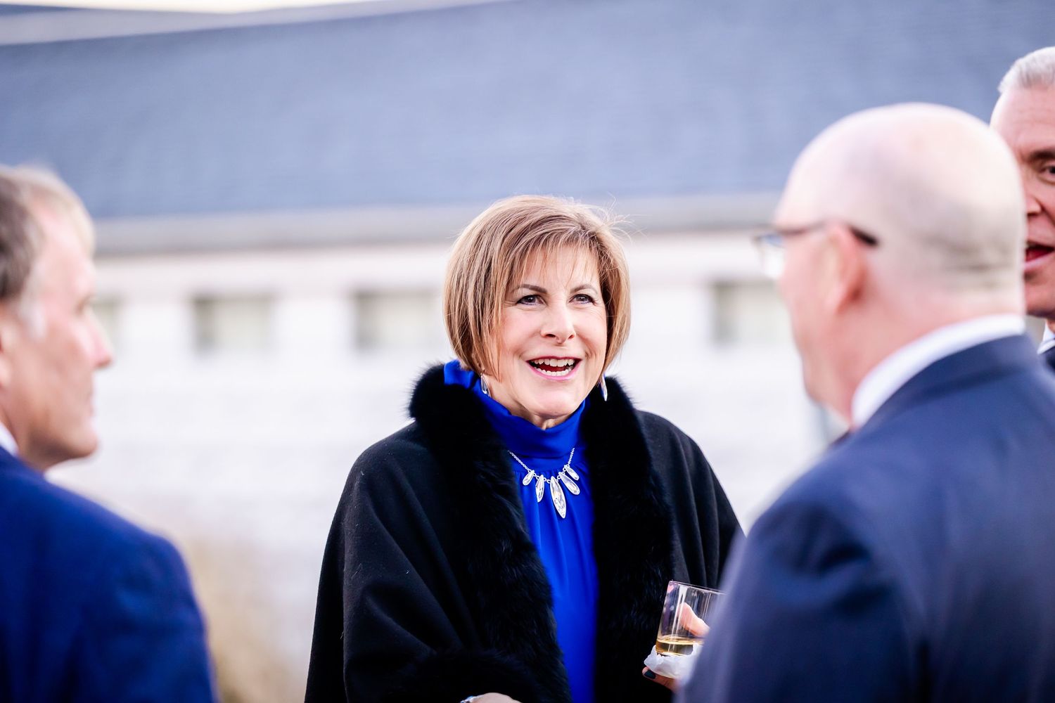 Woman in blue outfit and black cape smiling at an outdoor social gathering with other guests.