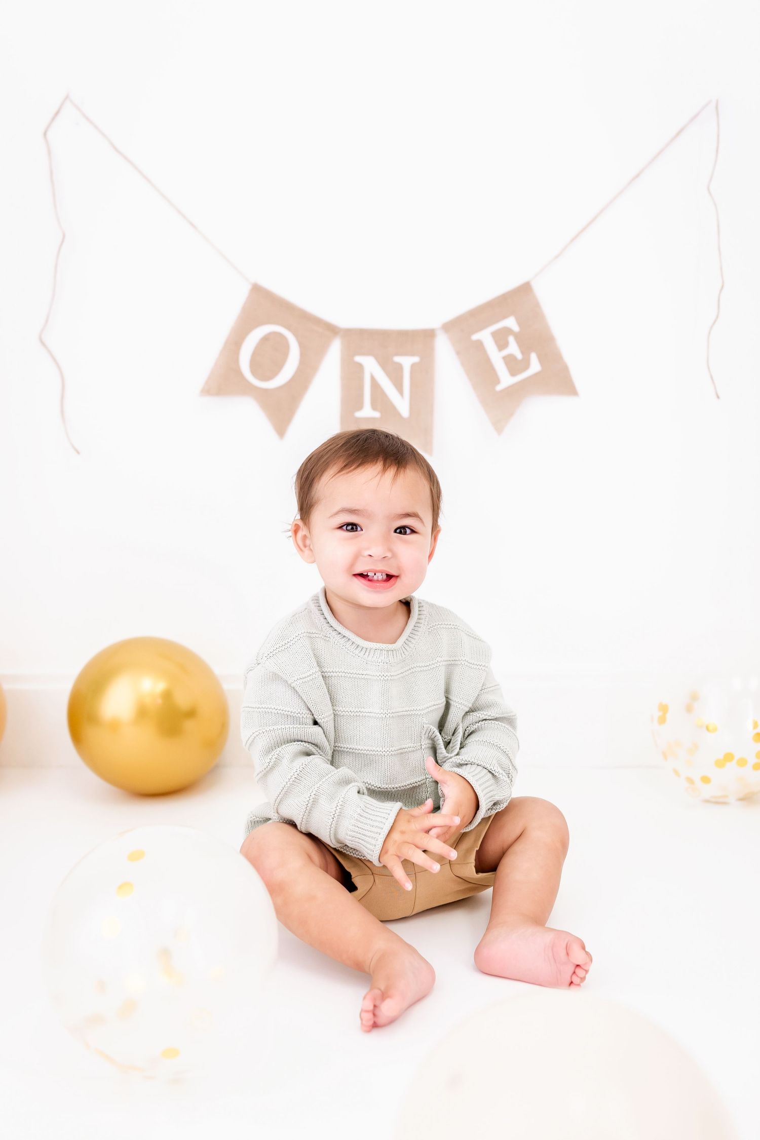Baby in gray sweater sitting under 'ONE' banner with gold balloon for first birthday celebration.