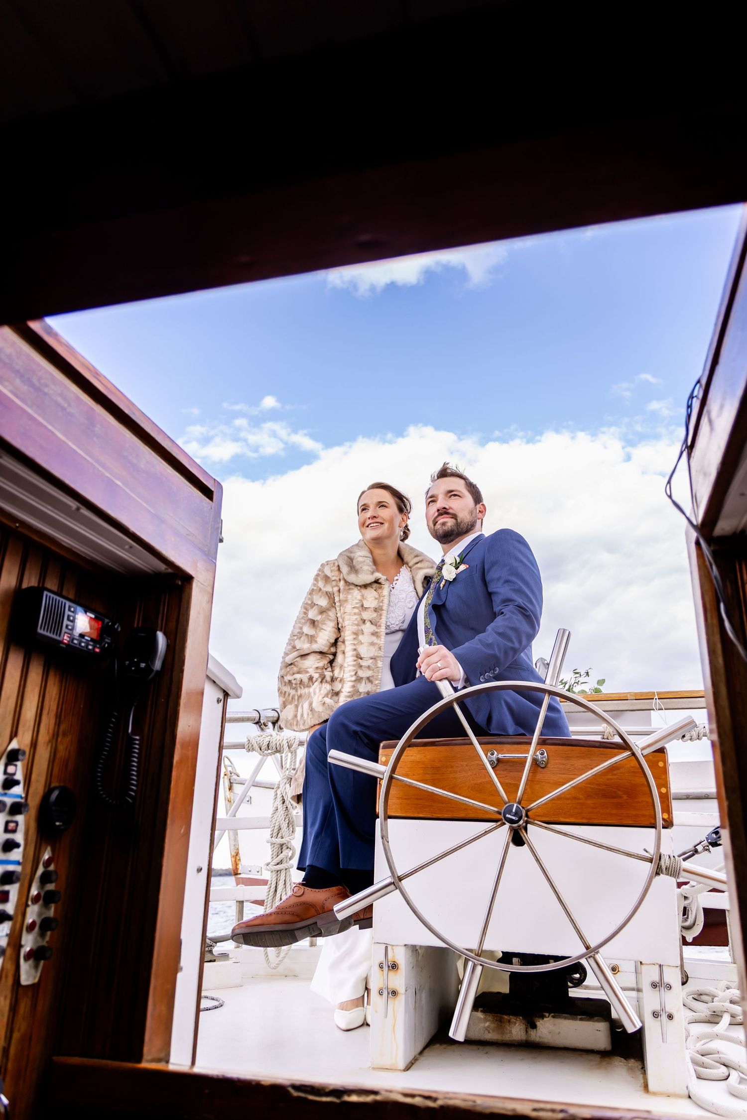 Two people enjoying a sunny day at the helm of a sailboat, with one wearing a fur coat and another in a blue suit.