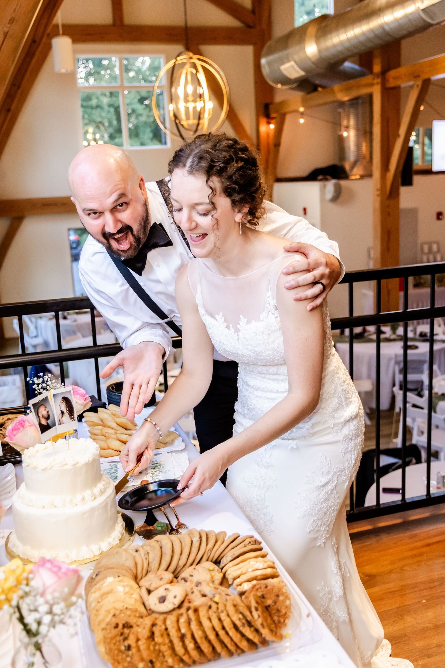 A couple in wedding attire cuts their wedding cake together in a rustic wooden venue with natural lighting.