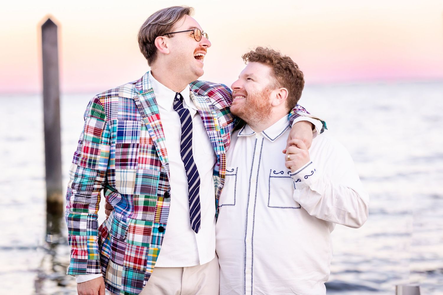 Two people in coordinating outfits laugh together while walking on a beach at sunset.