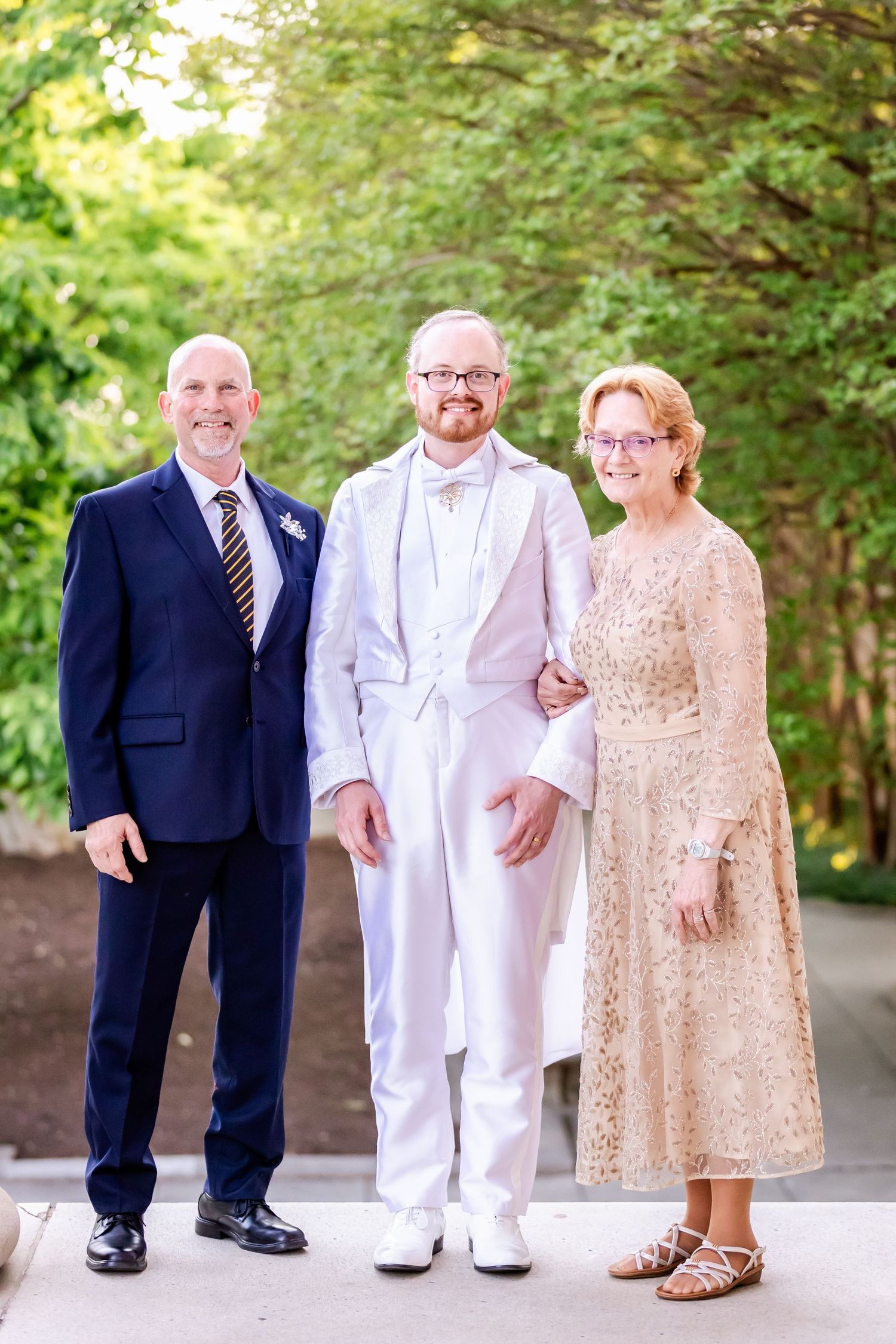 Three wedding party members pose together with one in white and two in navy and champagne formal wear.