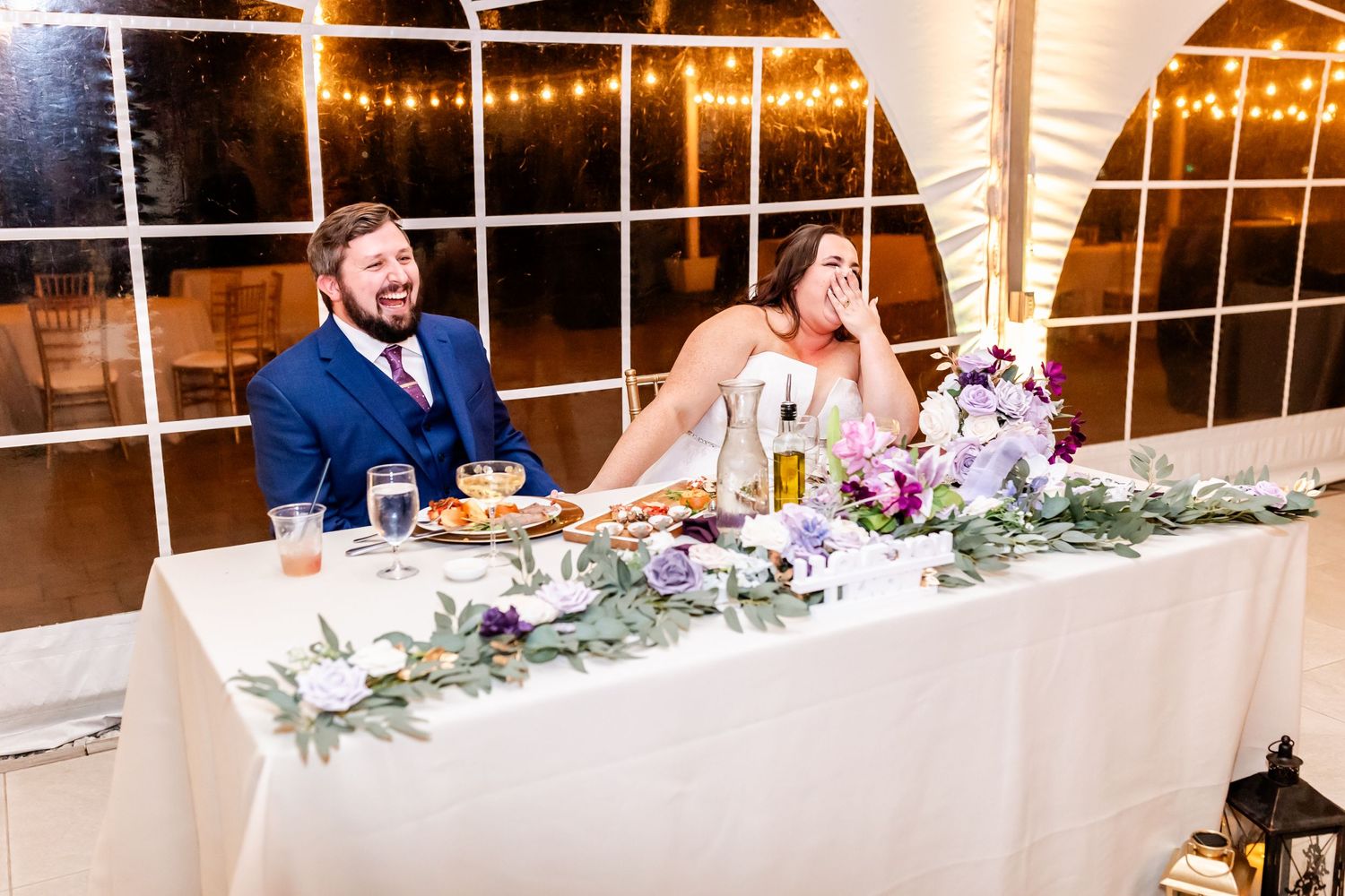 Couple enjoying their wedding reception at decorated head table with floral arrangements.