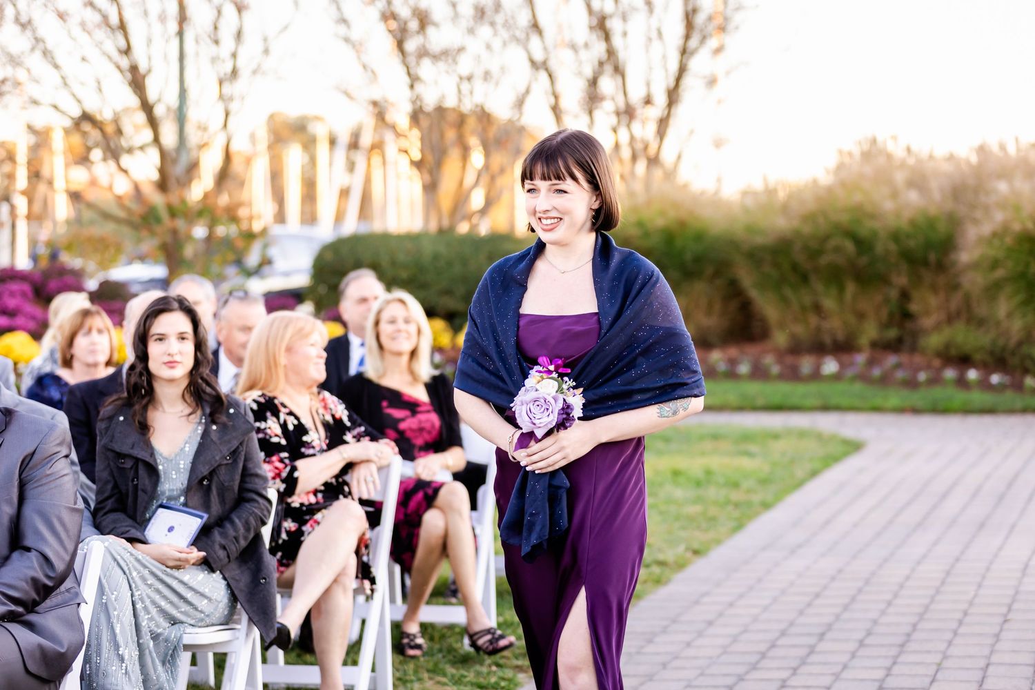 Wedding party member in purple dress and shawl walking down outdoor ceremony aisle.