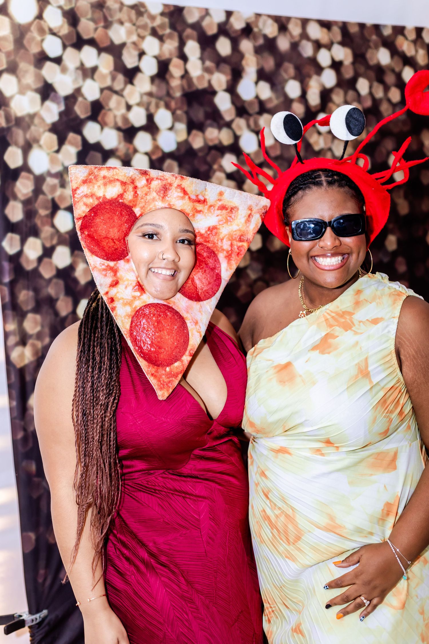 Two party guests wearing festive pizza and crab themed costumes pose against sparkly backdrop.