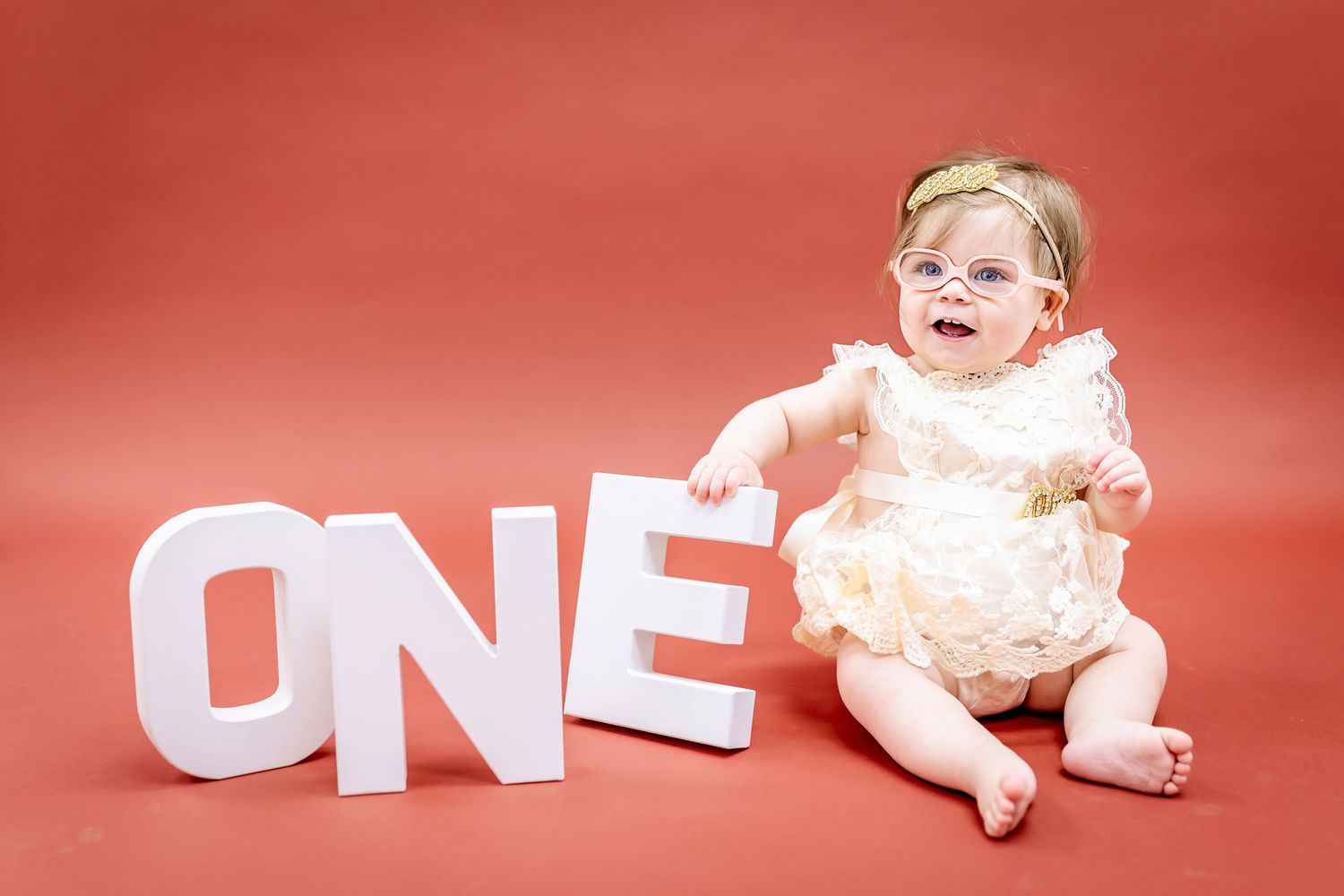 A baby in cream dress sits beside white block letters spelling 'ONE' on a red background.