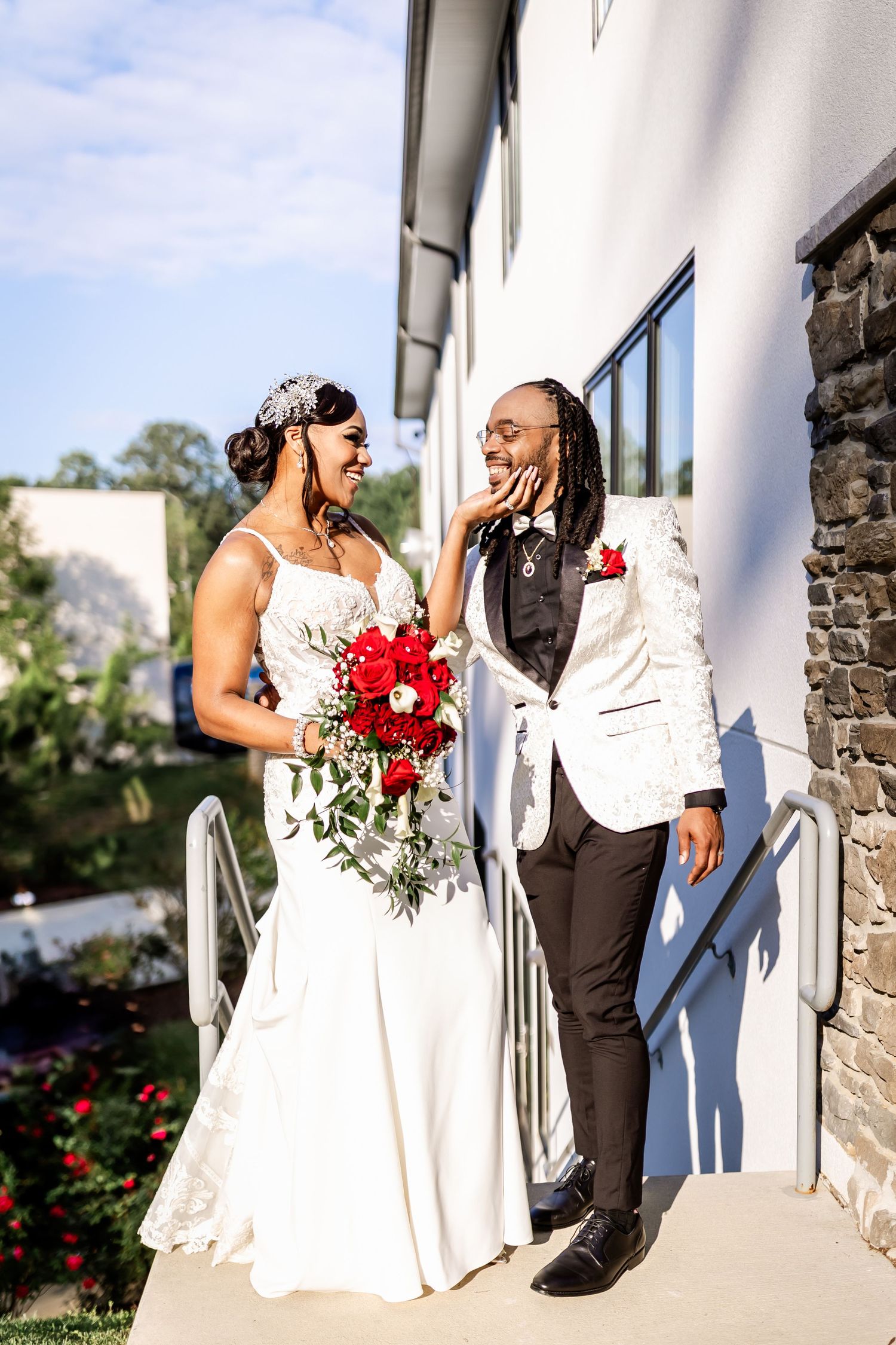 Bride and groom share joyful moment on outdoor terrace with red rose bouquet.