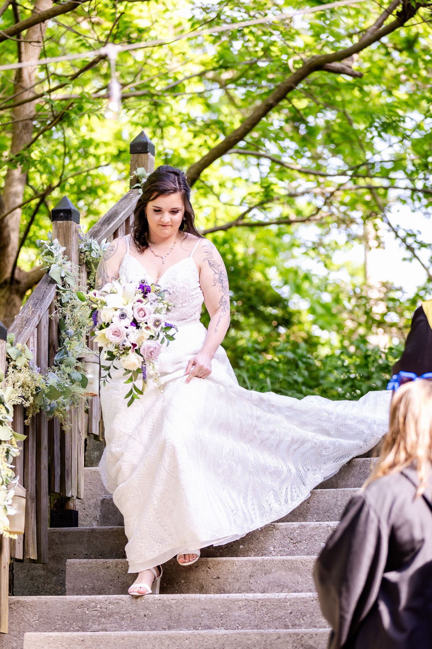 A bride in a white lace dress descends stone stairs while carrying a purple and white floral bouquet.