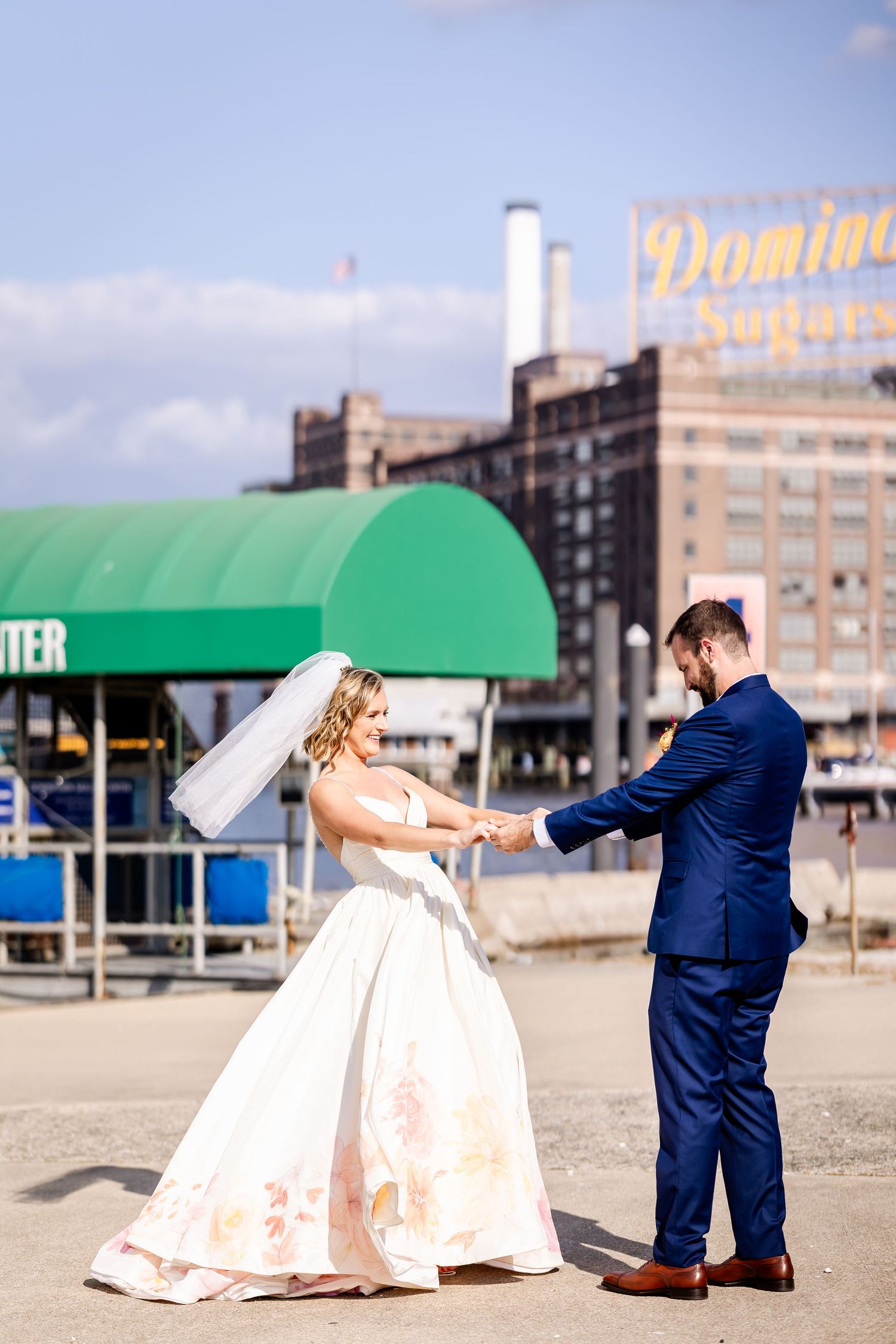 A couple dances and twirls in front of a waterfront industrial backdrop with green awning and Domino sign visible.