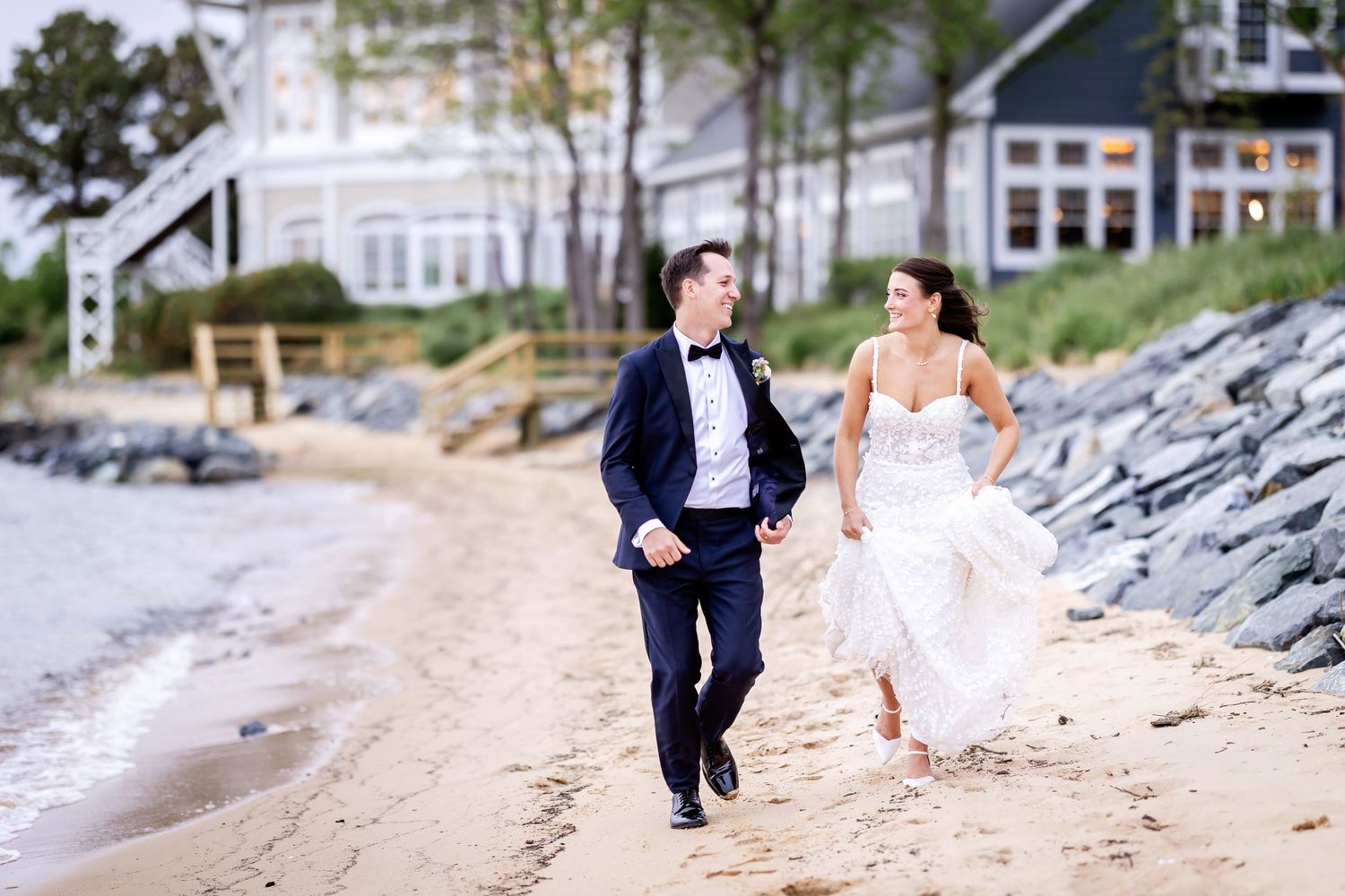 Newlywed couple runs joyfully along a sandy beach with a coastal house and rocky shoreline in the background.