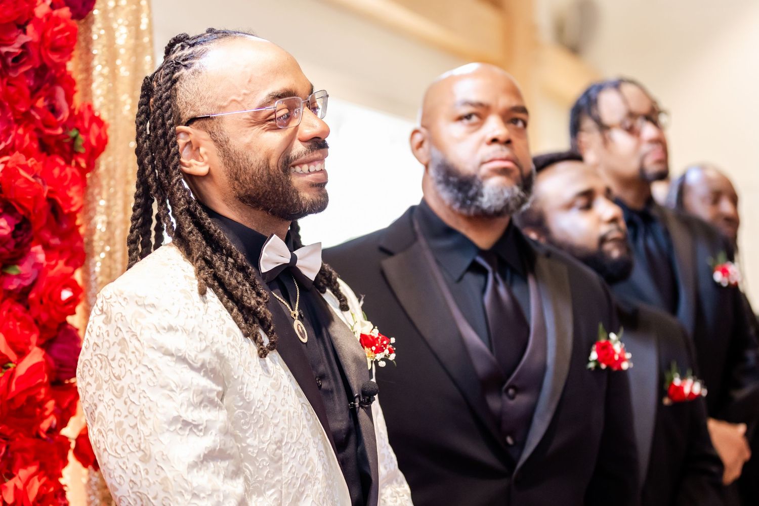 Groomsmen in formal attire stand near red floral display during wedding ceremony.