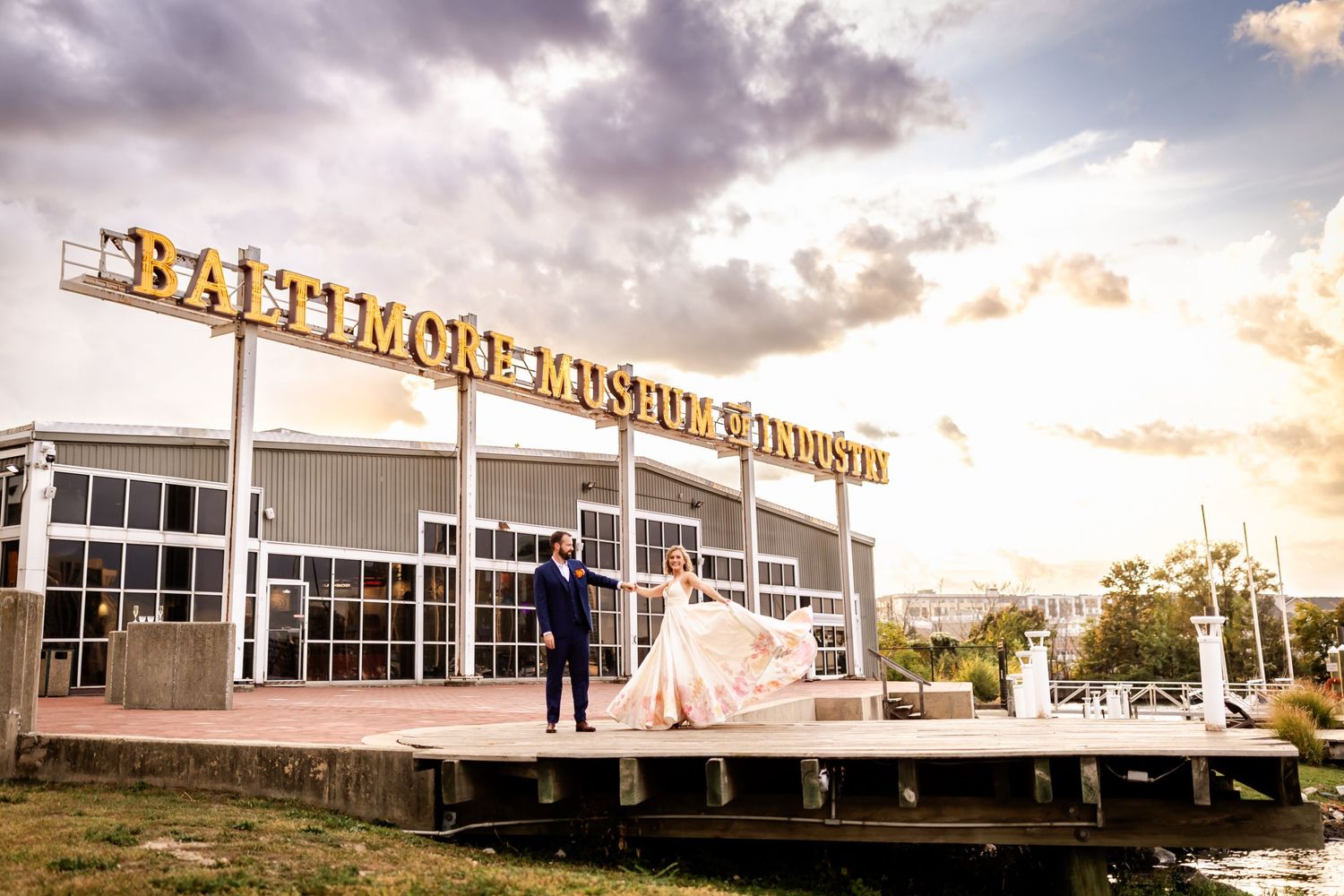 A couple in wedding attire stands on a platform against a sunset sky with illuminated vintage building signage.