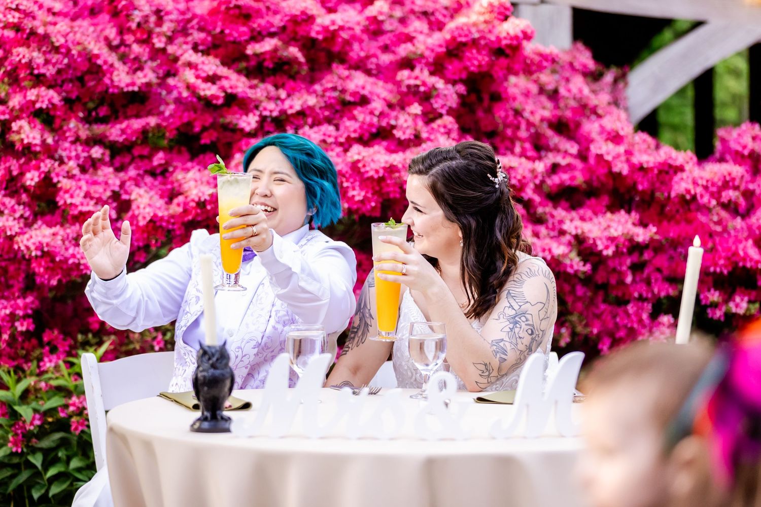 Wedding guests laugh together at reception table with vibrant pink azalea bushes in background.