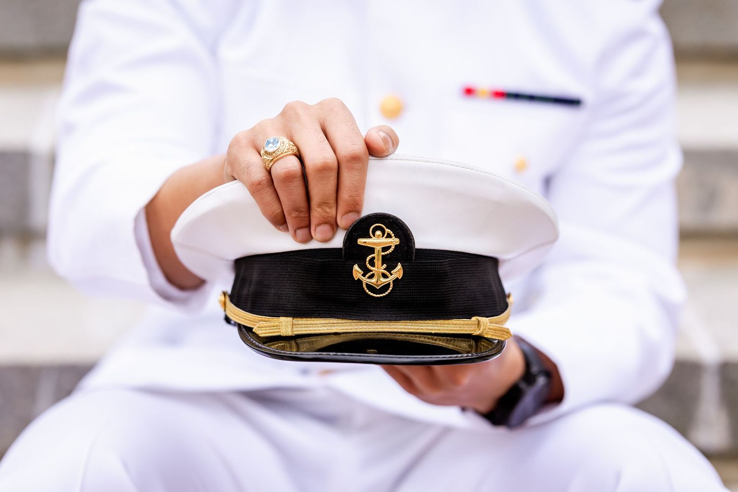 Hands in white uniform hold a naval officer's cap with gold braid and anchor emblem against blurred background.