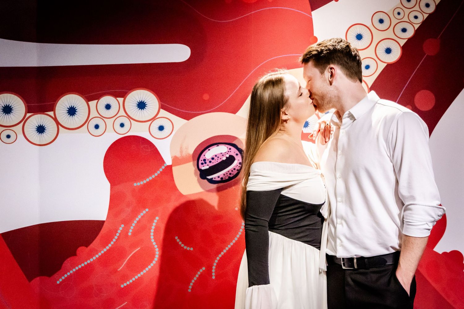A couple shares an intimate moment against a vibrant red wall with circular artistic patterns.