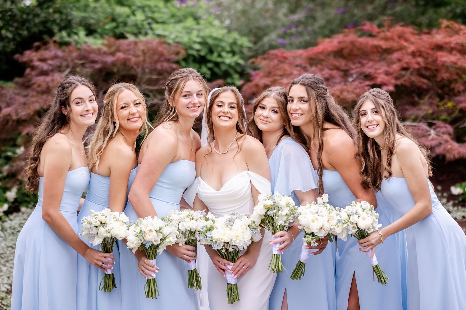 Bridal party in light blue dresses poses with bouquets against vibrant red maple trees.