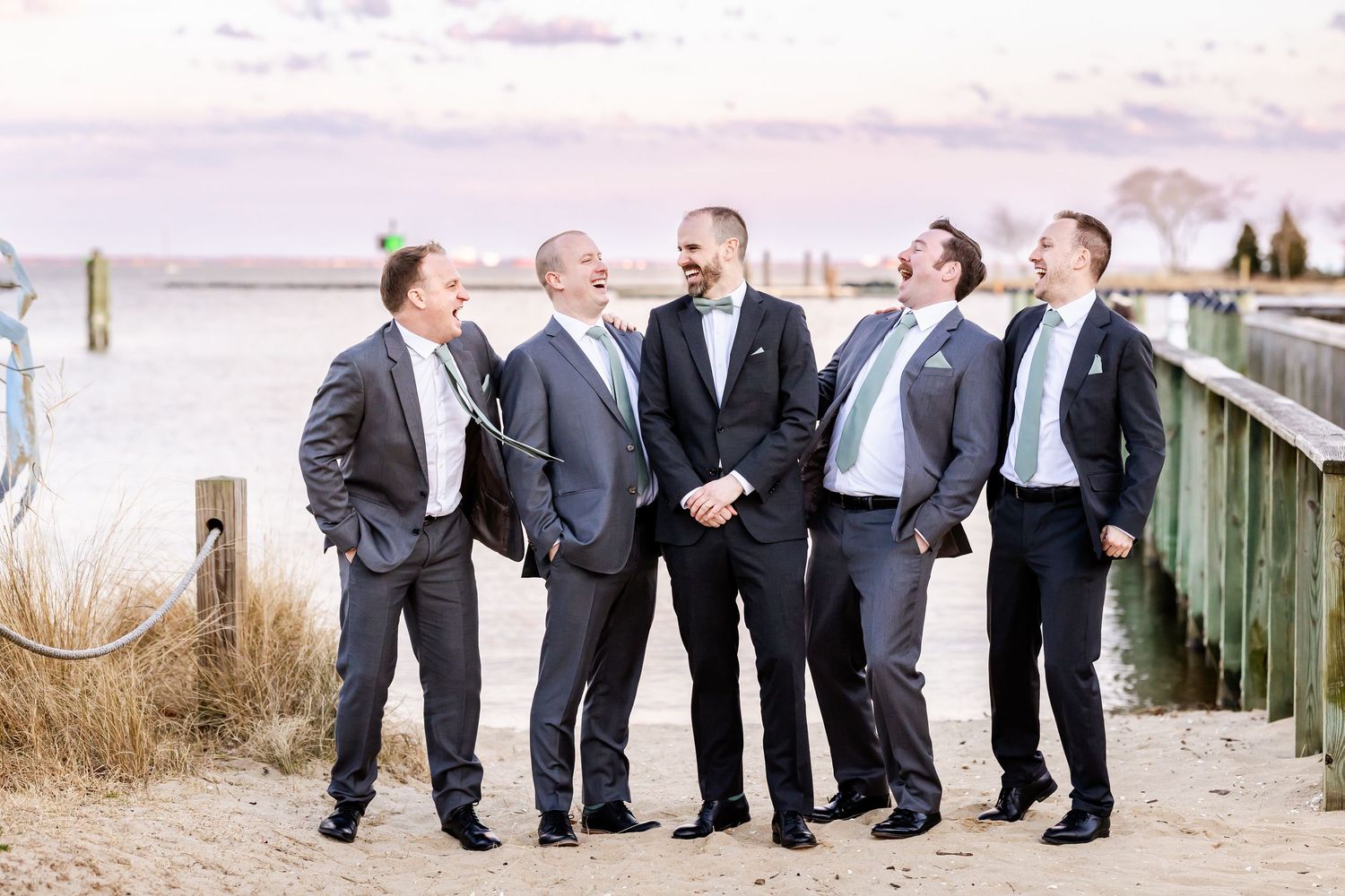 Groomsmen in dark suits stand in a candid line along a wooden boardwalk at an outdoor venue.