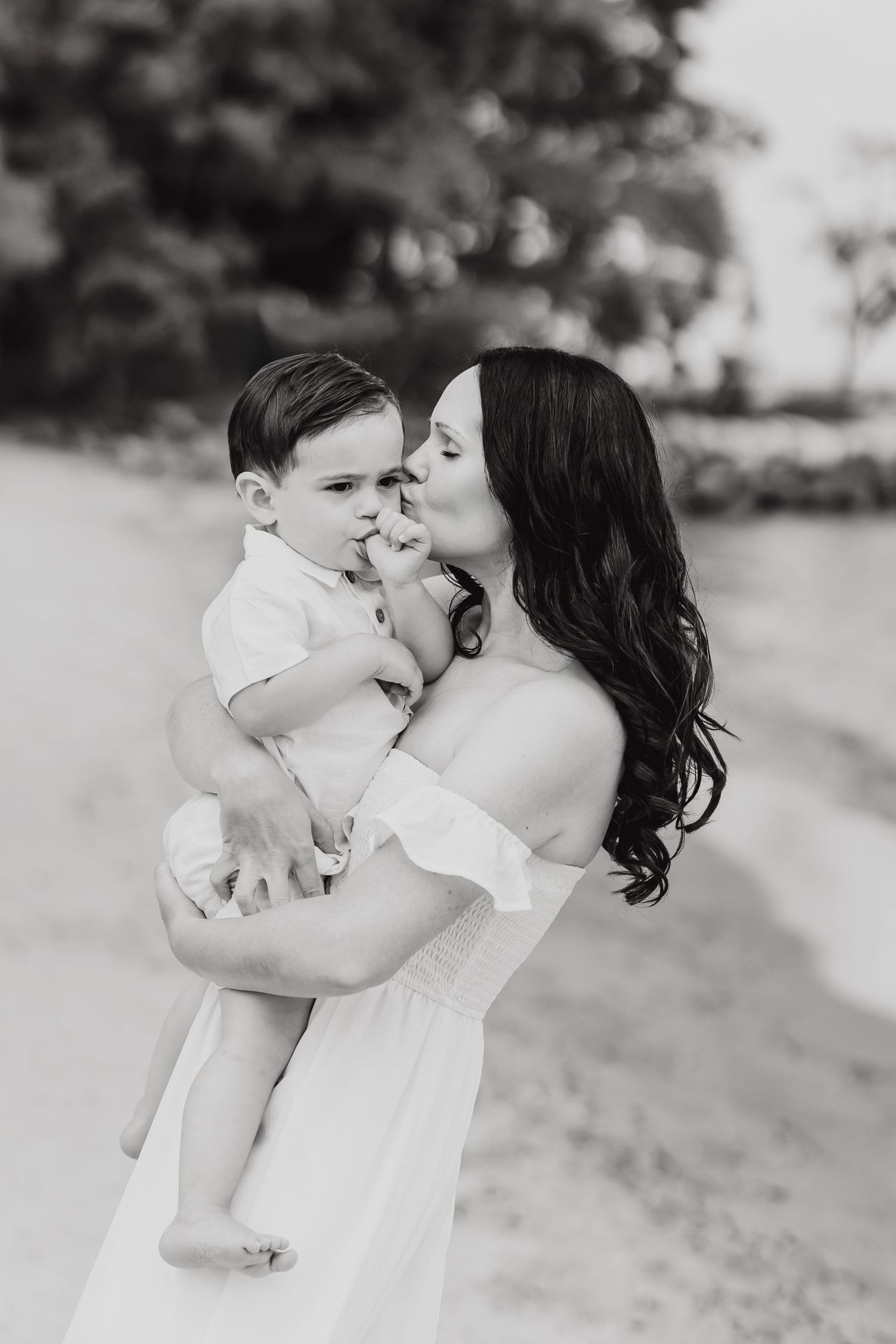 Black and white portrait of woman in off-shoulder dress holding toddler outdoors.