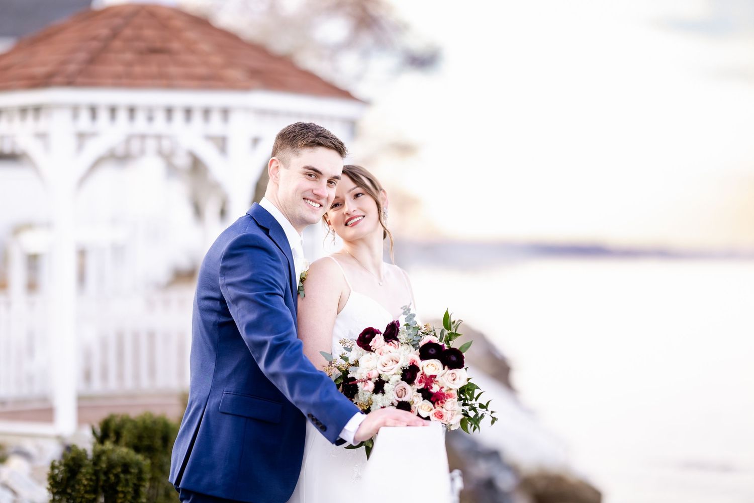 Romantic wedding couple embraces on a white balcony overlooking the water at sunset.