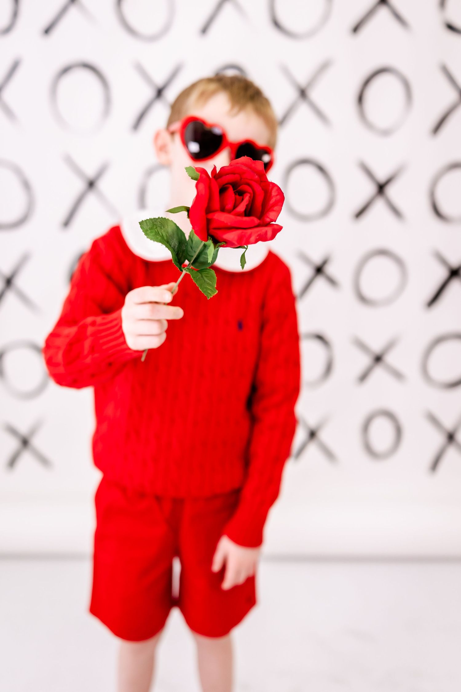 A young person in a bright red outfit holding a red rose while wearing heart-shaped sunglasses against an XOXO backdrop.