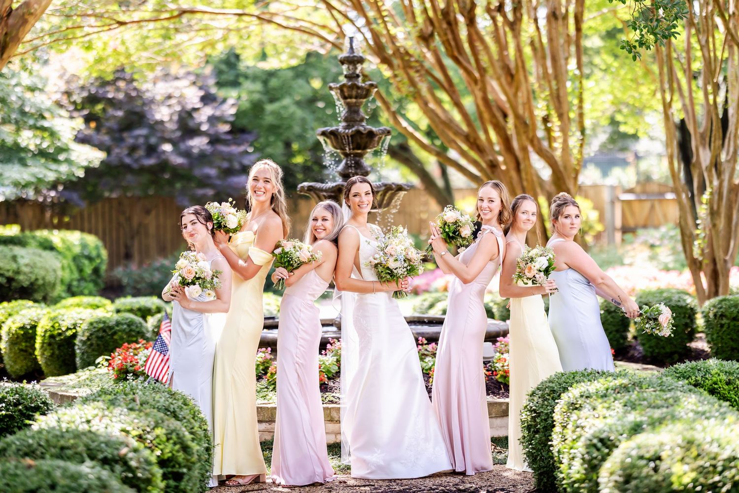 A wedding party poses together by a garden fountain surrounded by manicured hedges and trees.
