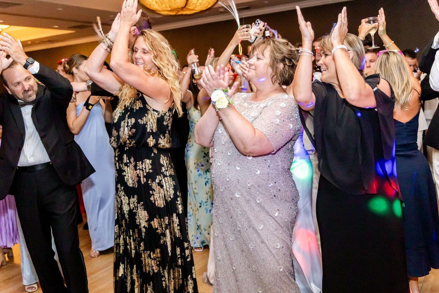 Wedding party guests dancing with hands raised on a crowded dance floor under chandeliers.