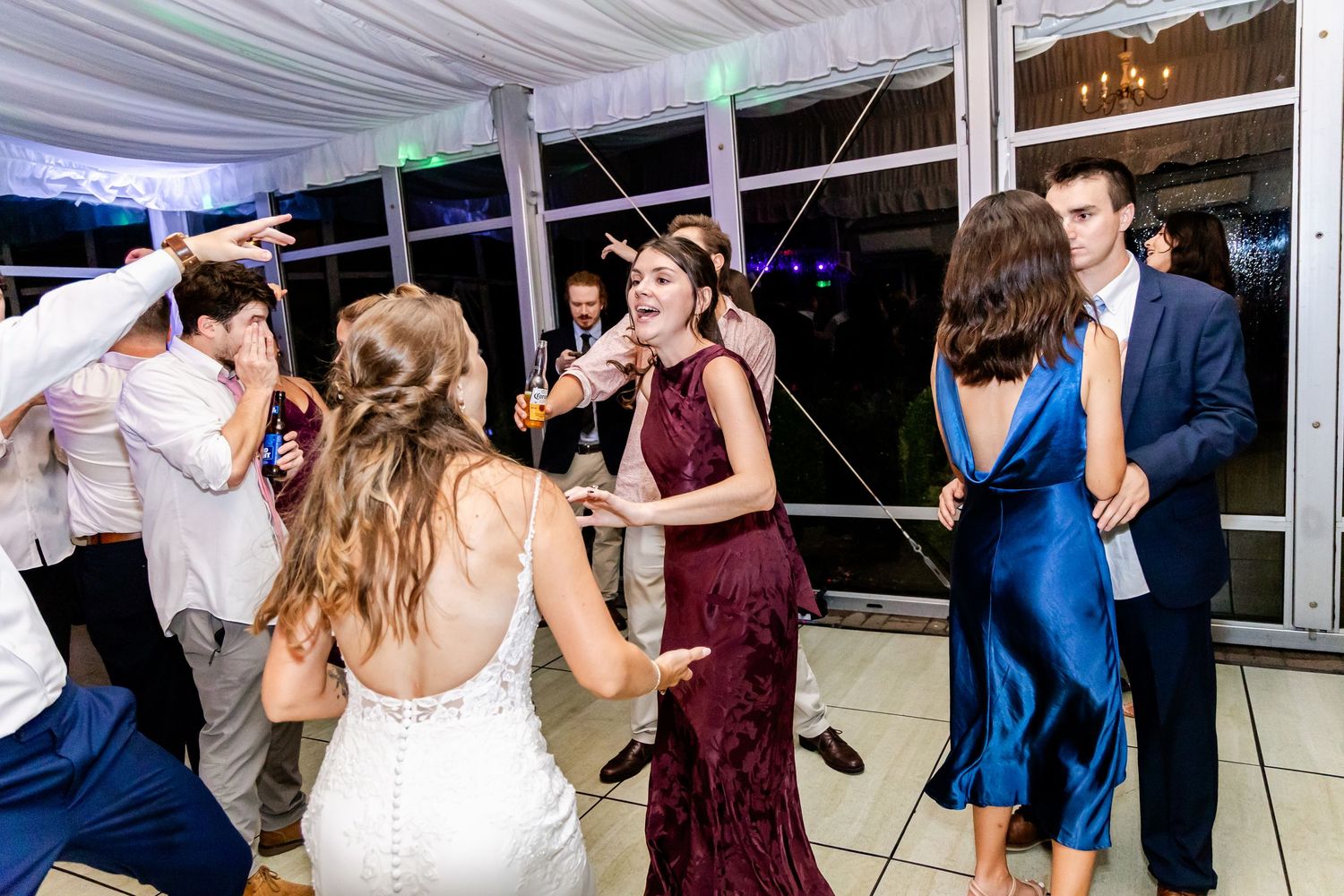 Wedding party dancing and celebrating at reception under tent with colorful lighting.
