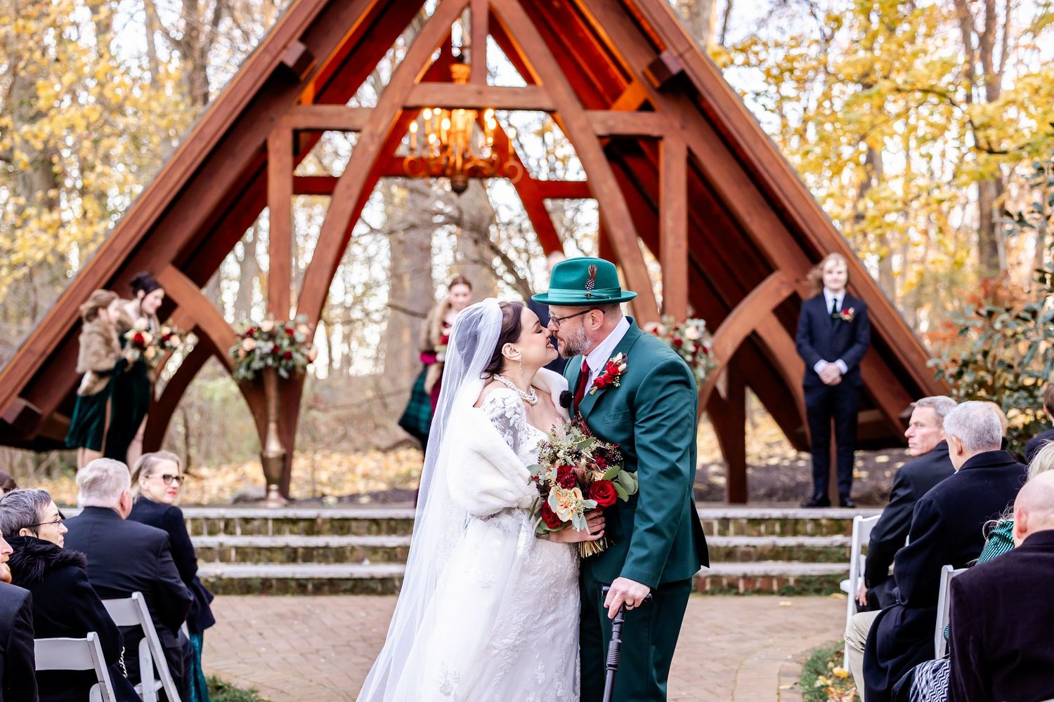 A wedding ceremony at a rustic outdoor chapel with wooden beams and fall foliage.