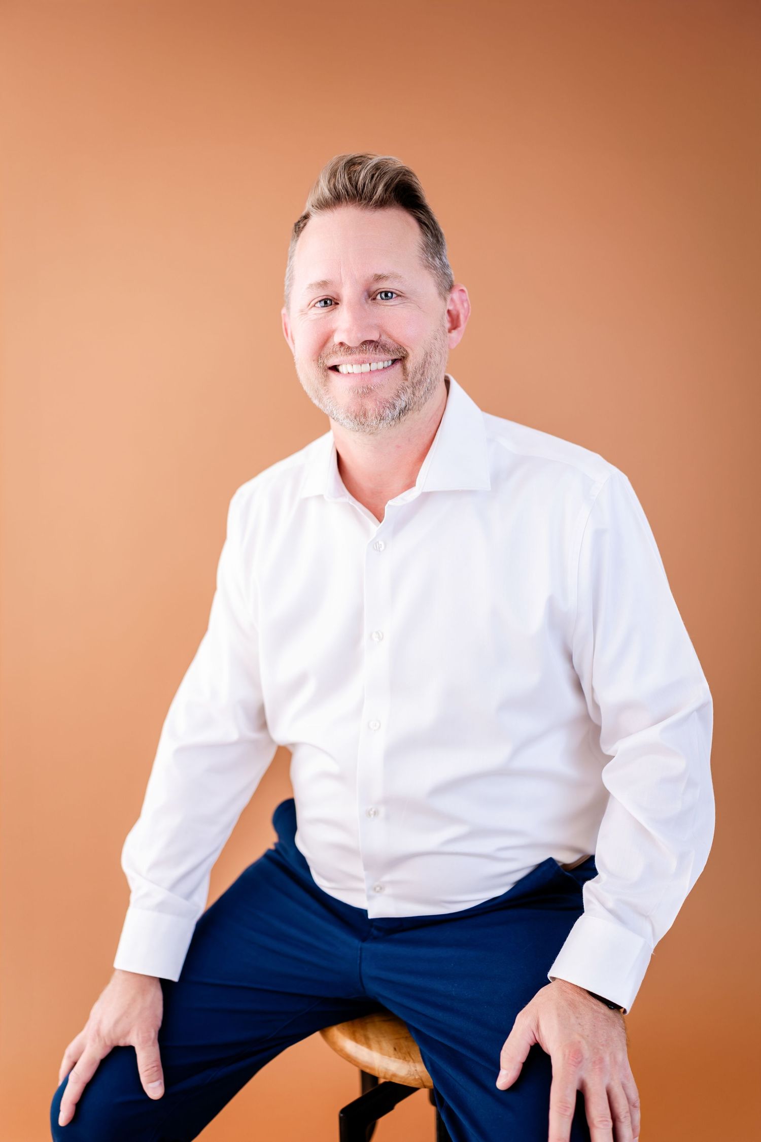 Smiling middle-aged man in white shirt and blue pants sitting on stool against warm orange background.
