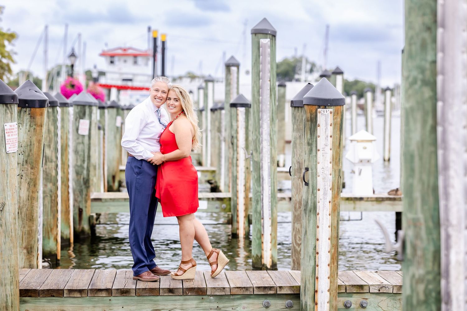 Couple poses together on a marina boardwalk with boats and buildings in the background.