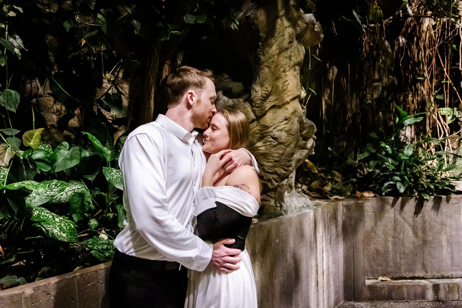 A romantic couple embraces in front of a lush garden wall with dramatic lighting and greenery.
