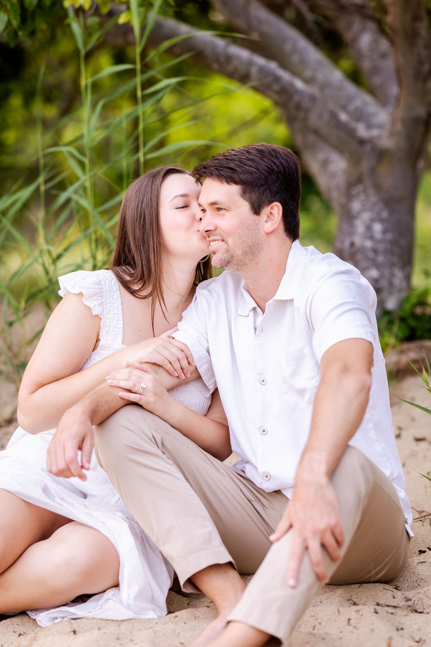 A couple in white clothing sharing a romantic moment while sitting together on sandy ground near lush foliage.