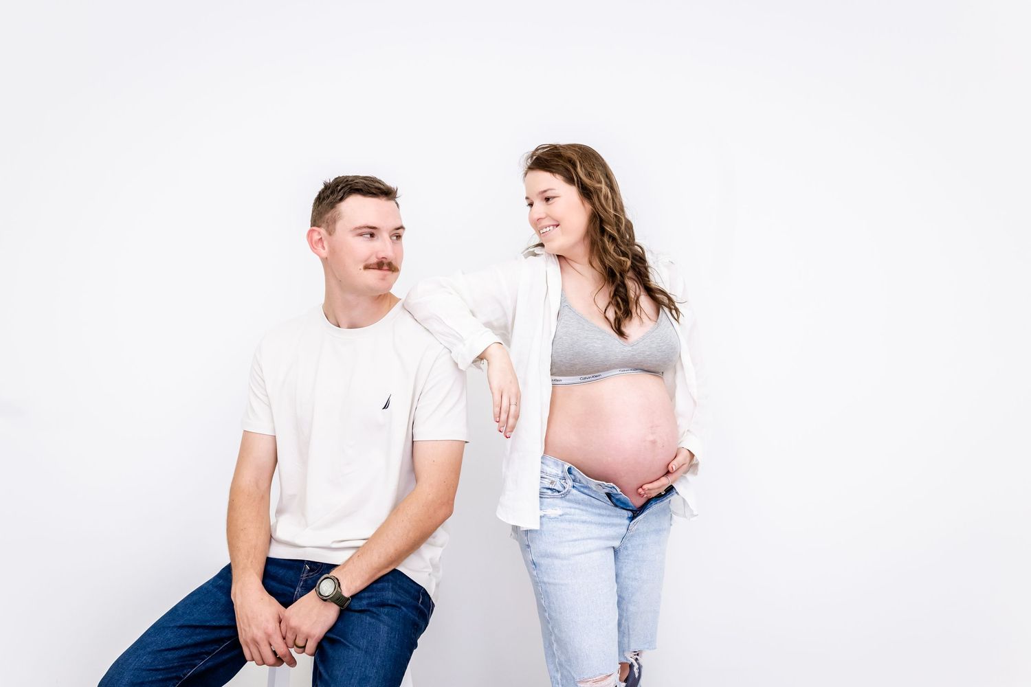 A couple wearing casual attire poses together against a white background during a maternity photoshoot.