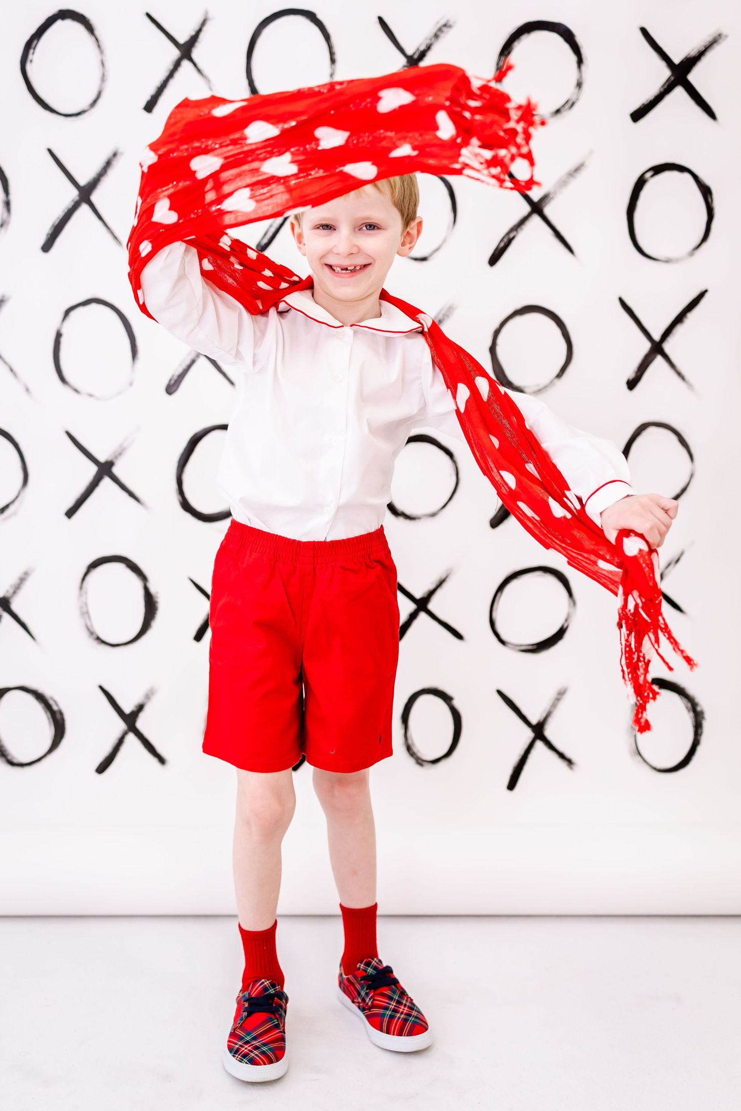 A child in red shorts and white shirt stands against a black and white X's and O's background with flowing red fabric.