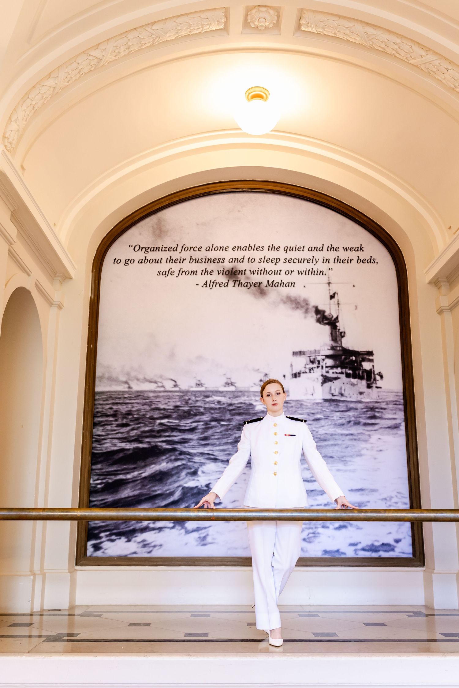 A white-clad figure stands in front of a naval-themed display featuring a historical warship photograph and inspirational quote.