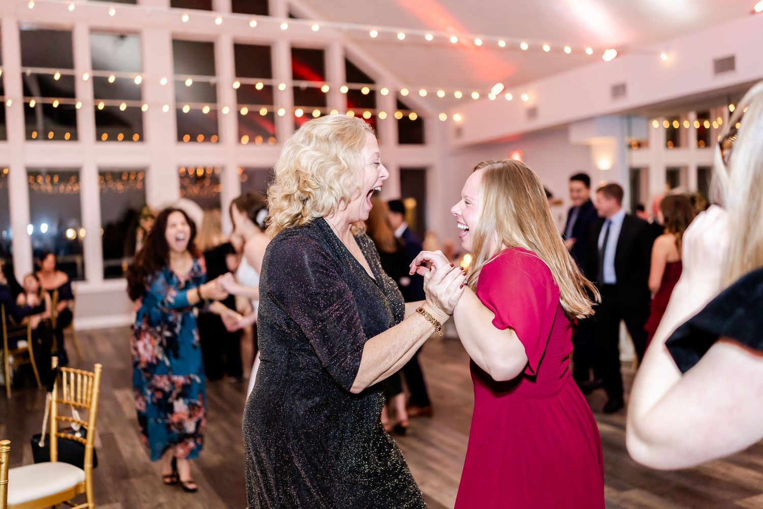 Wedding guests dancing together on a wooden dance floor under string lights at an indoor reception.