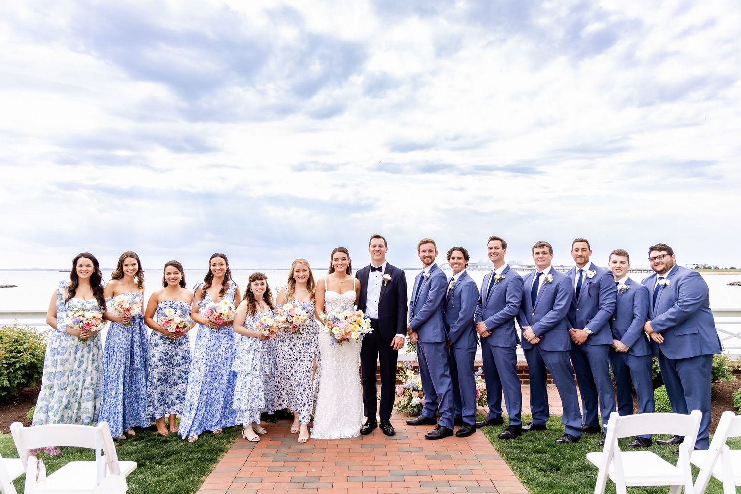 Large wedding party in blue suits and floral dresses posed together on waterfront patio.