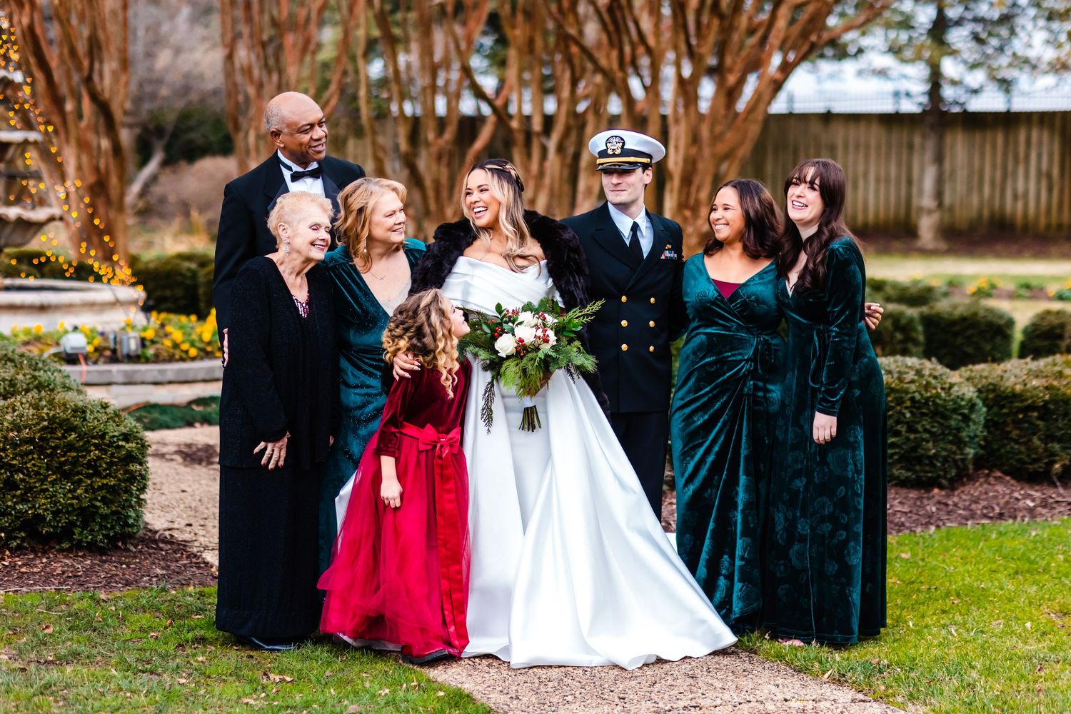 A wedding party gathers in a garden wearing formal attire including velvet gowns and navy military dress uniform.