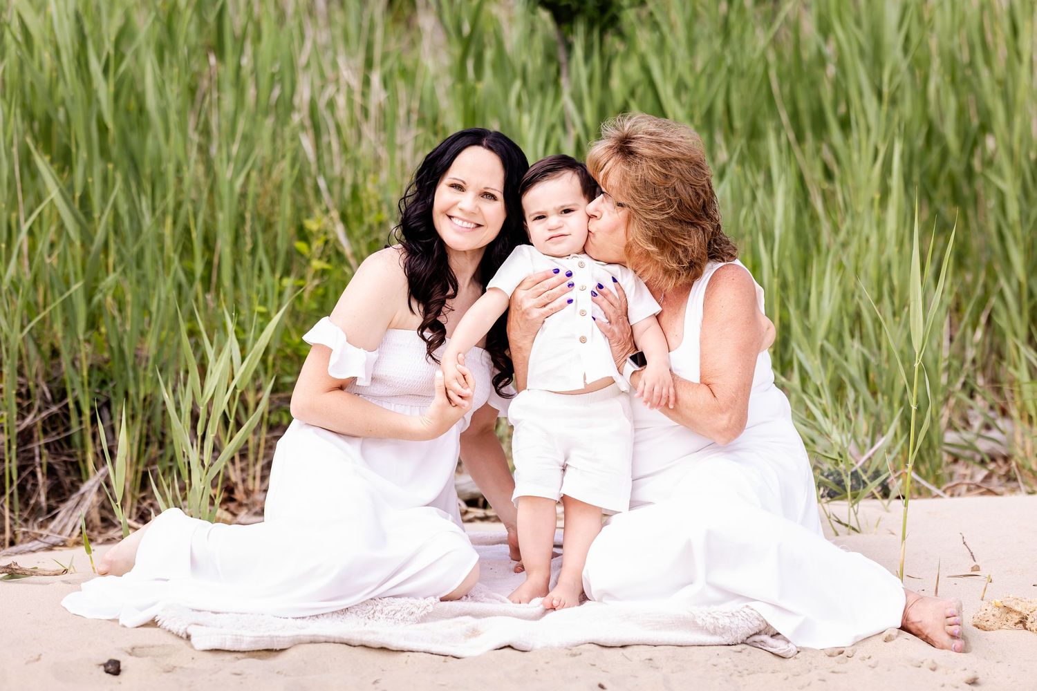 Family wearing white outfits sits together on sandy beach against tall grassy backdrop during summer photoshoot.