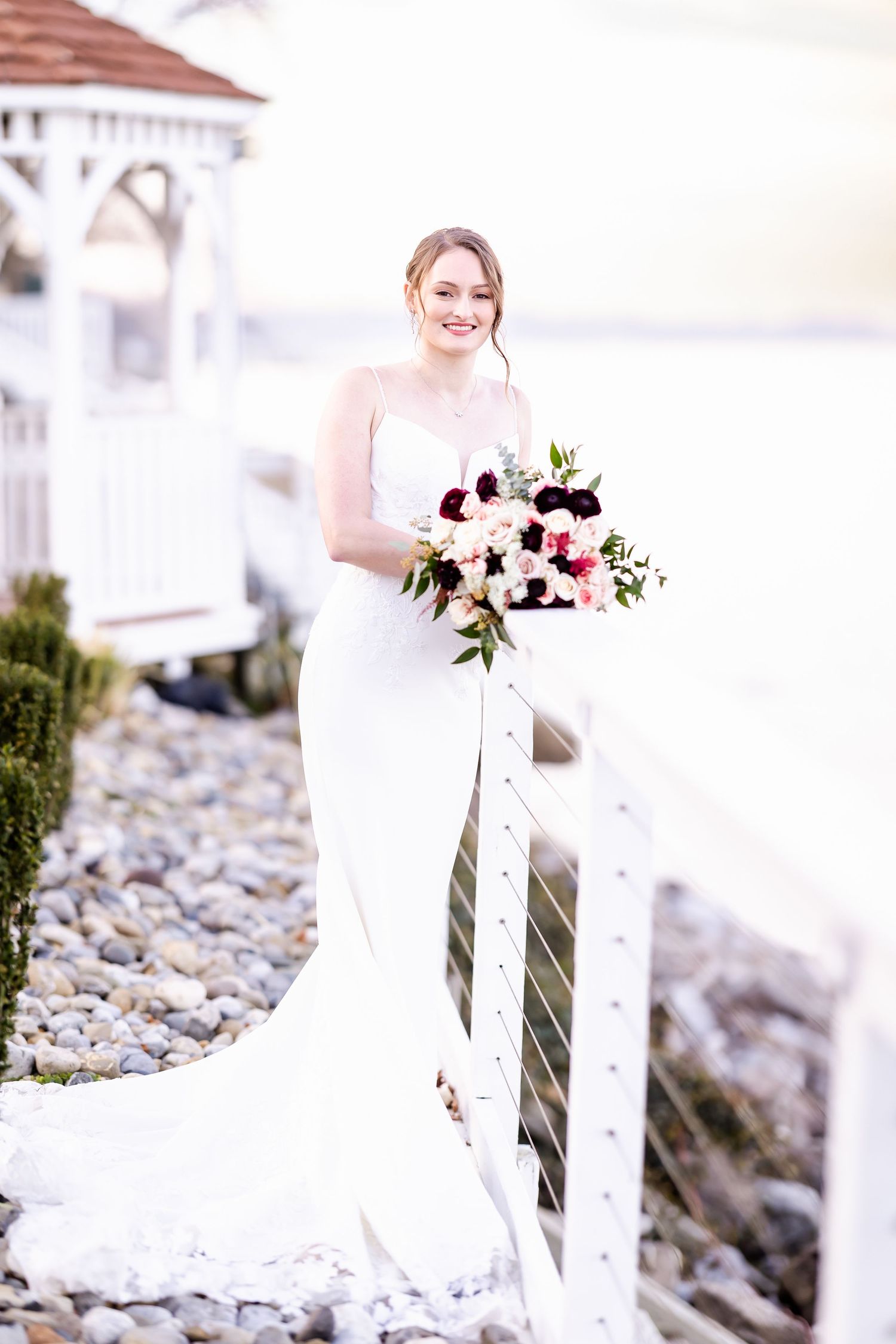 Bride in white dress holds bouquet while standing near rocky shoreline and white gazebo.
