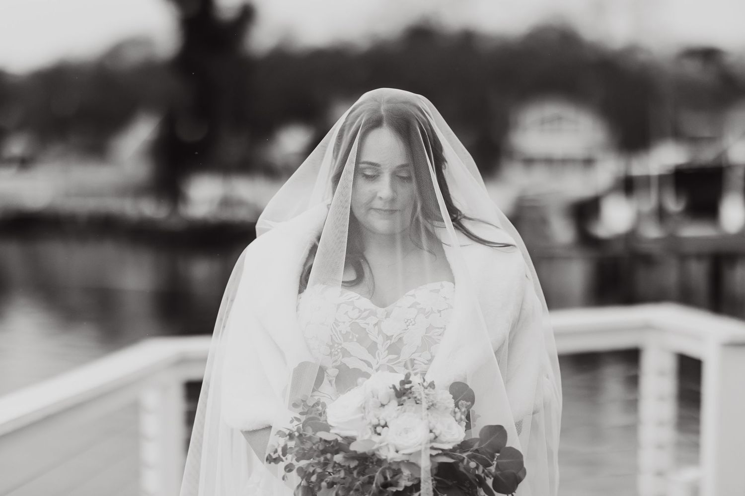 A black and white photo of a bride in a white veil and dress holding a floral bouquet while standing on a dock.