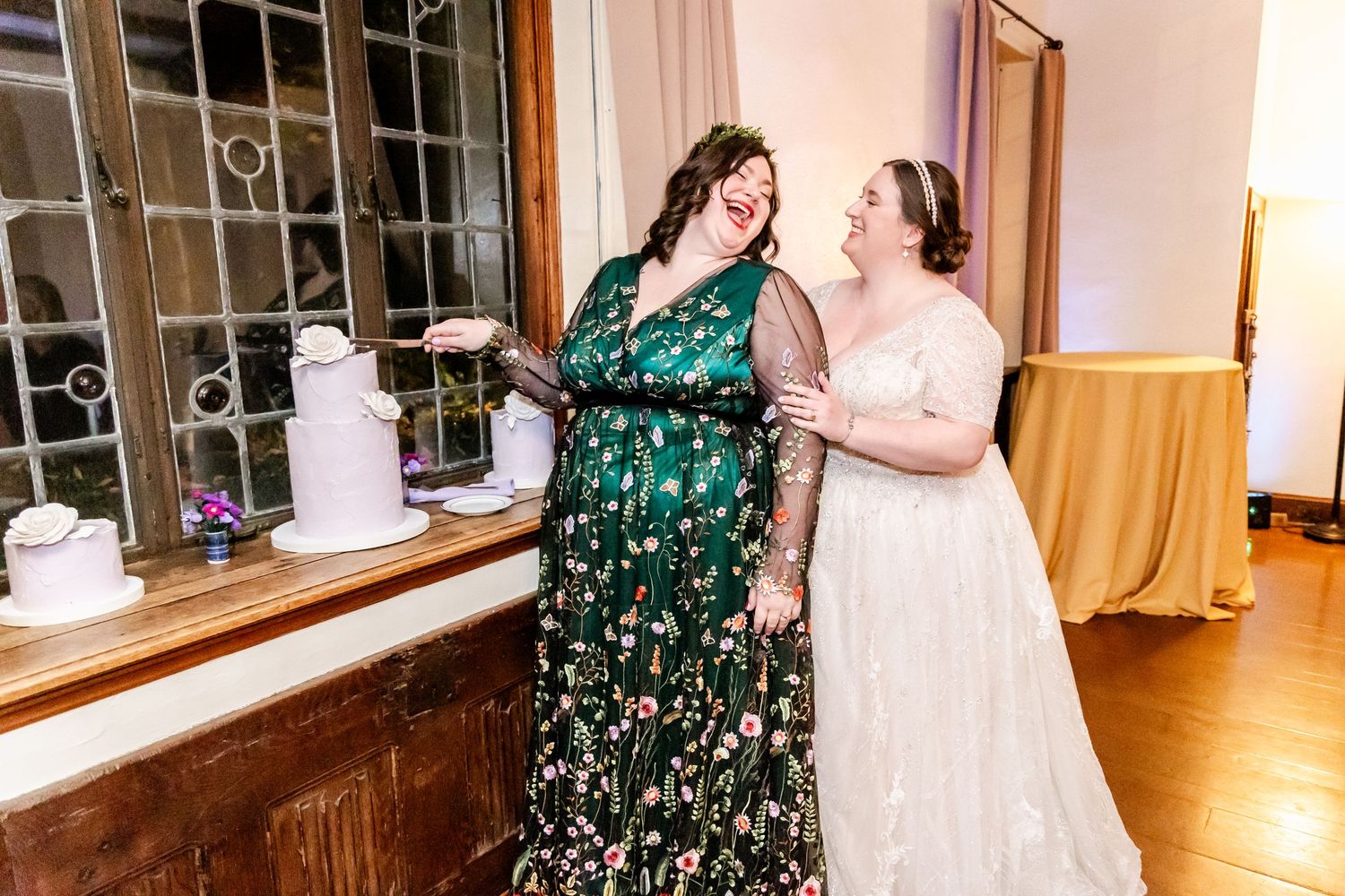 Two wedding guests in elegant dresses share a laughing moment at an indoor reception.