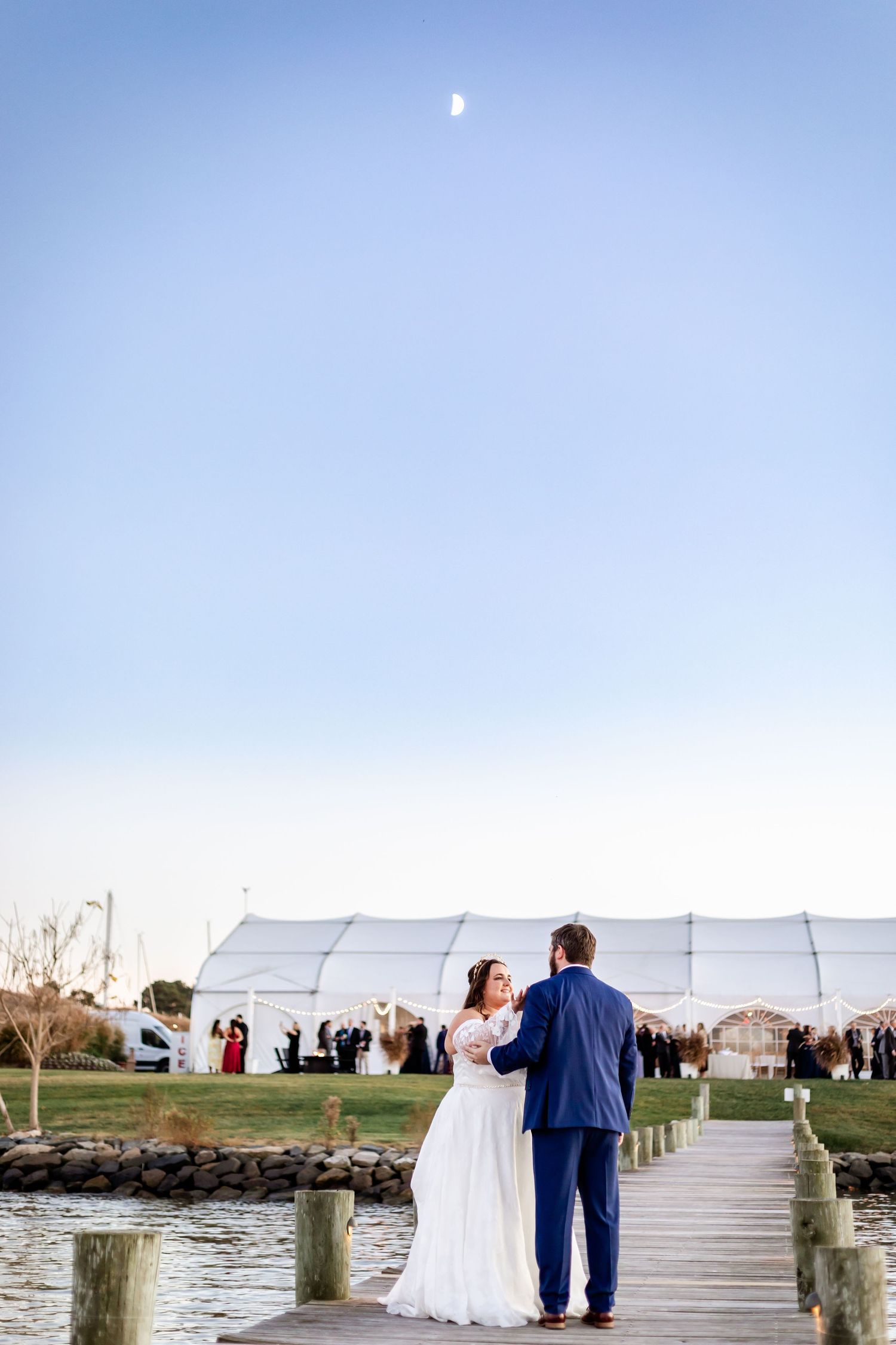 A couple stands on a waterfront dock with a white tent venue visible in the background.