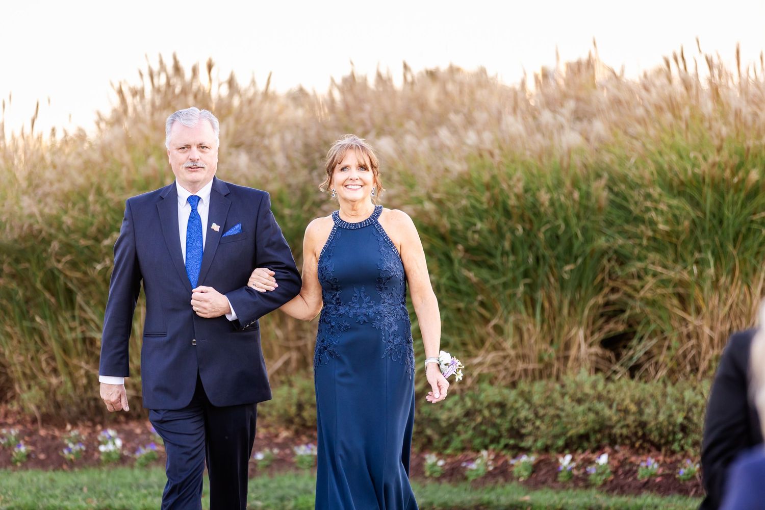 Formal couple walks arm in arm along garden path during evening event.