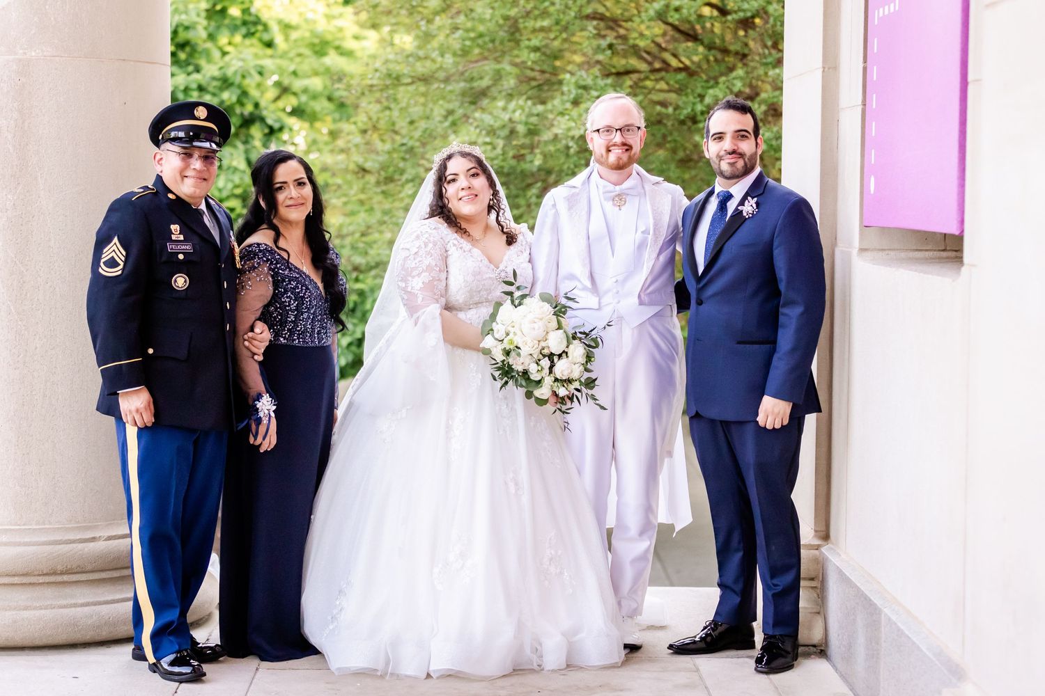 Wedding group portrait with military uniform and formal attire against garden backdrop.