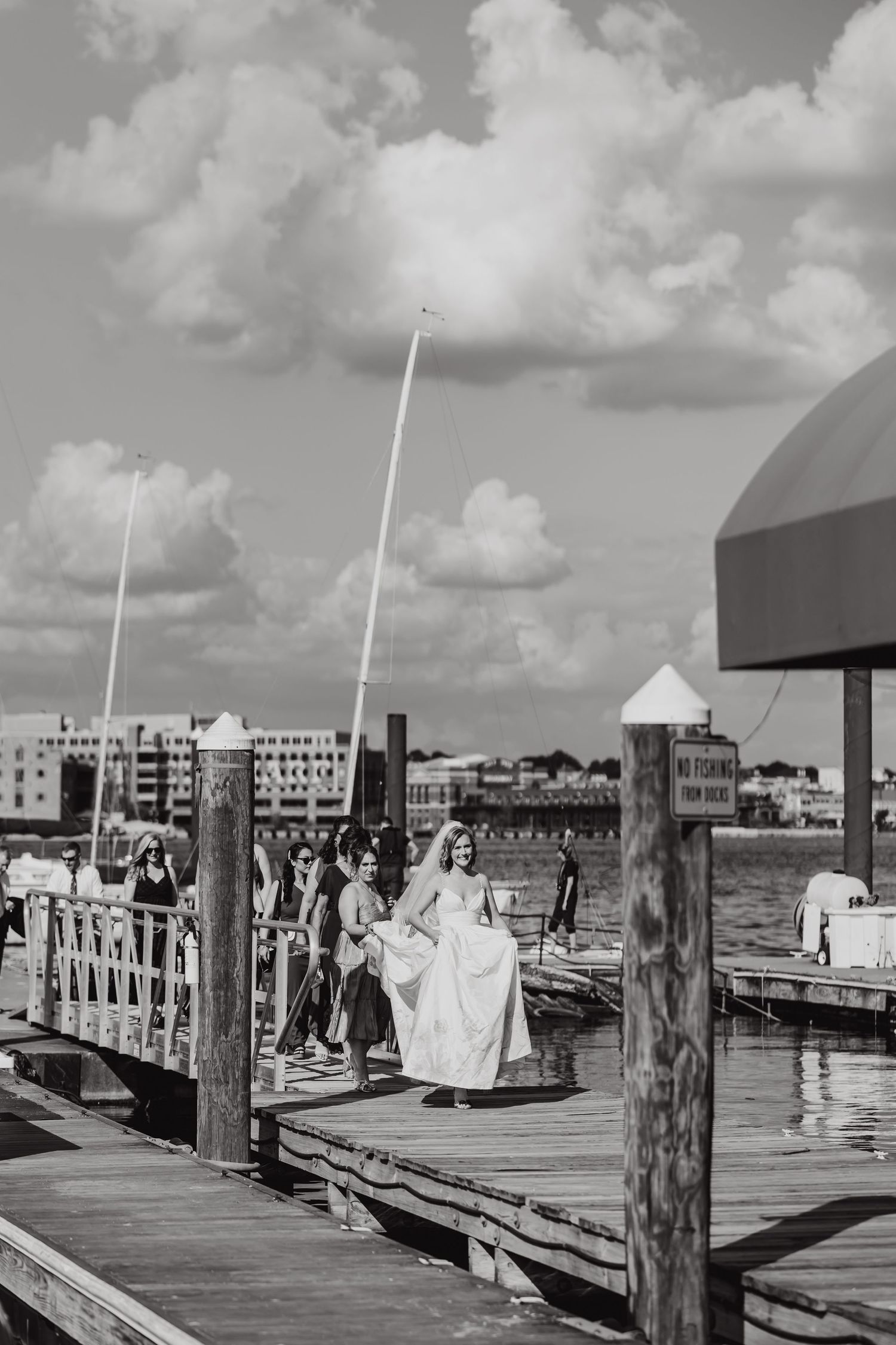 Black and white photo of a wedding party walking along a wooden dock by the water.
