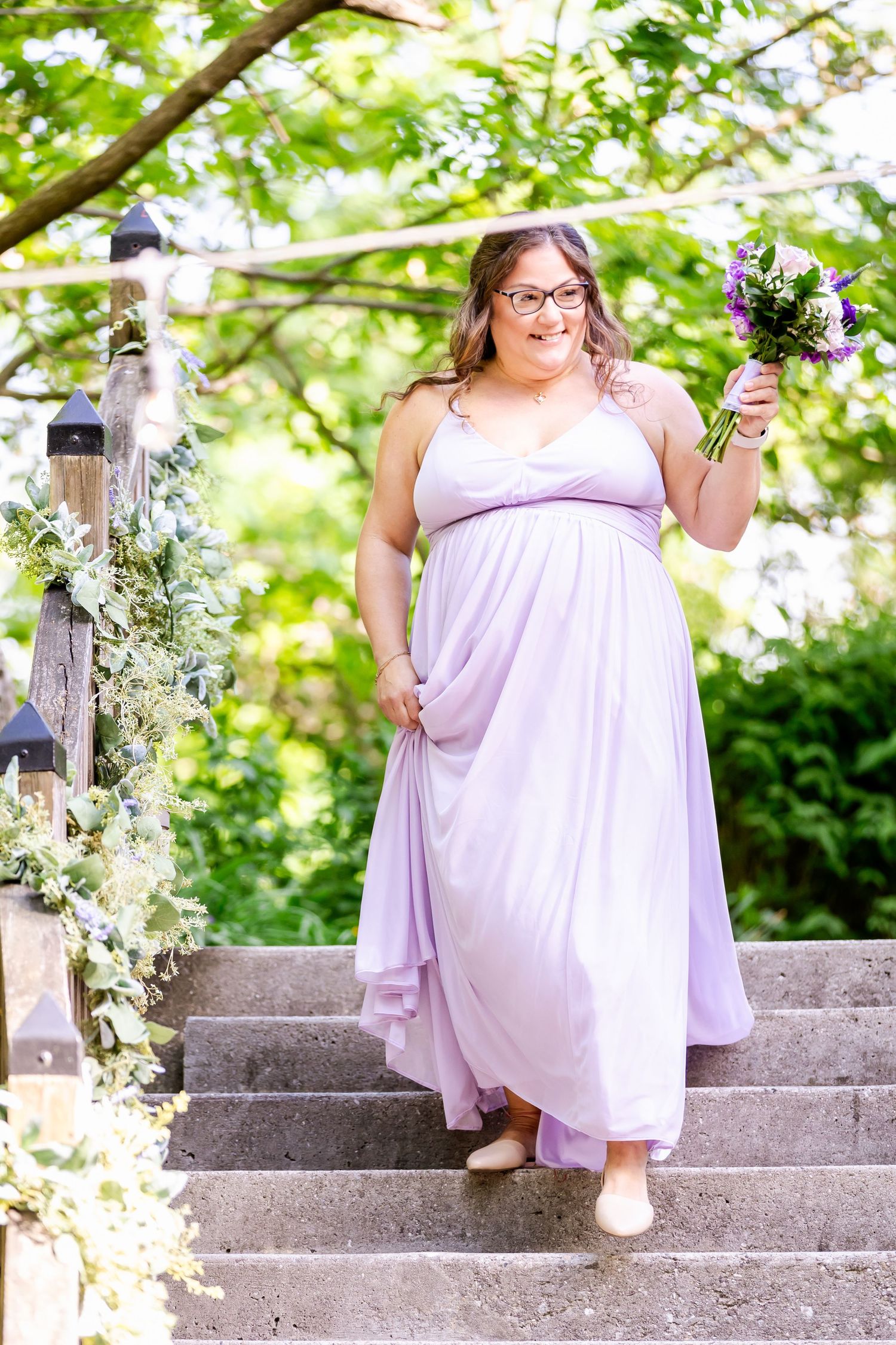 Woman in light purple dress holds bouquet while standing on stone steps decorated with flowers.