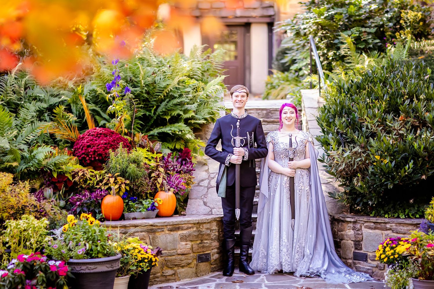 A couple stands in an autumn garden filled with colorful flowers and pumpkins on a stone patio during sunset.