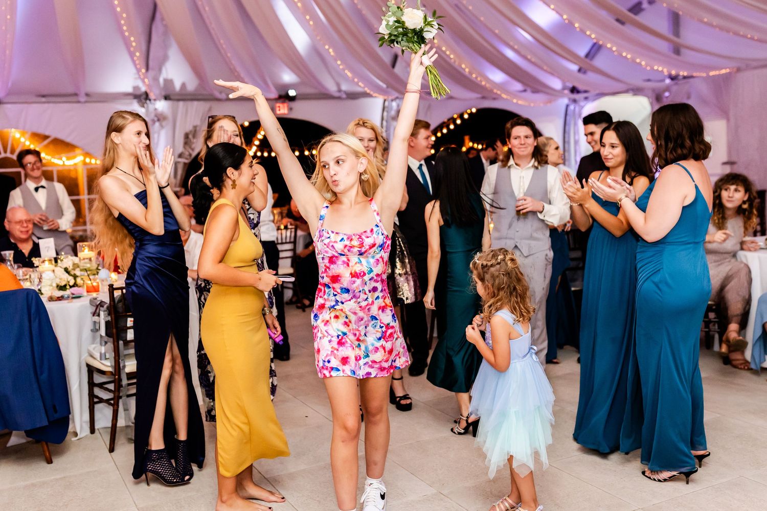People dancing and celebrating at a wedding reception inside a white tented venue with guests in colorful formal attire.